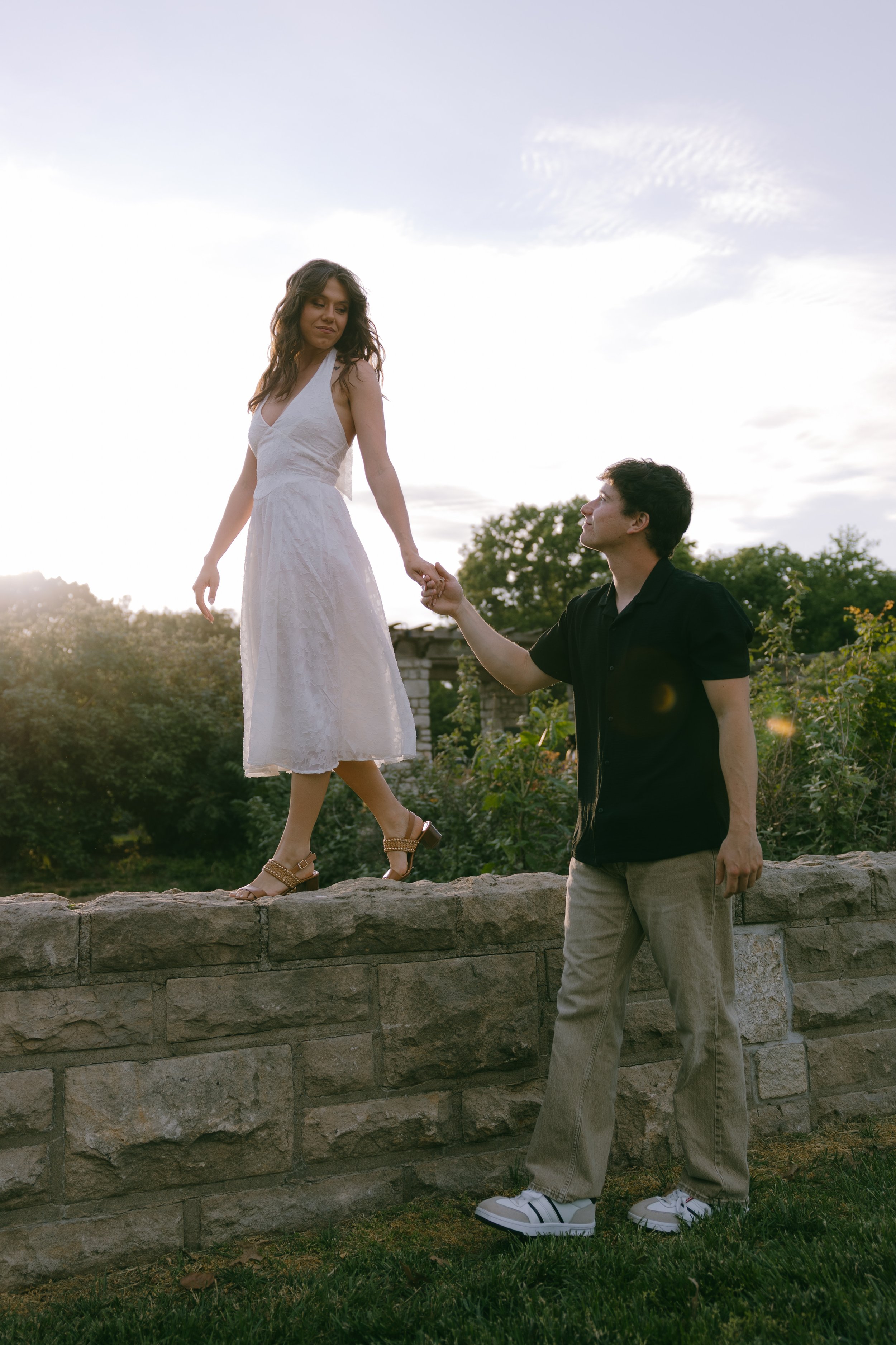 A man kneeling on a stone wall holding hands with a woman standing on the wall, in an outdoor setting with trees and a partly cloudy sky in the background during late afternoon or early evening.