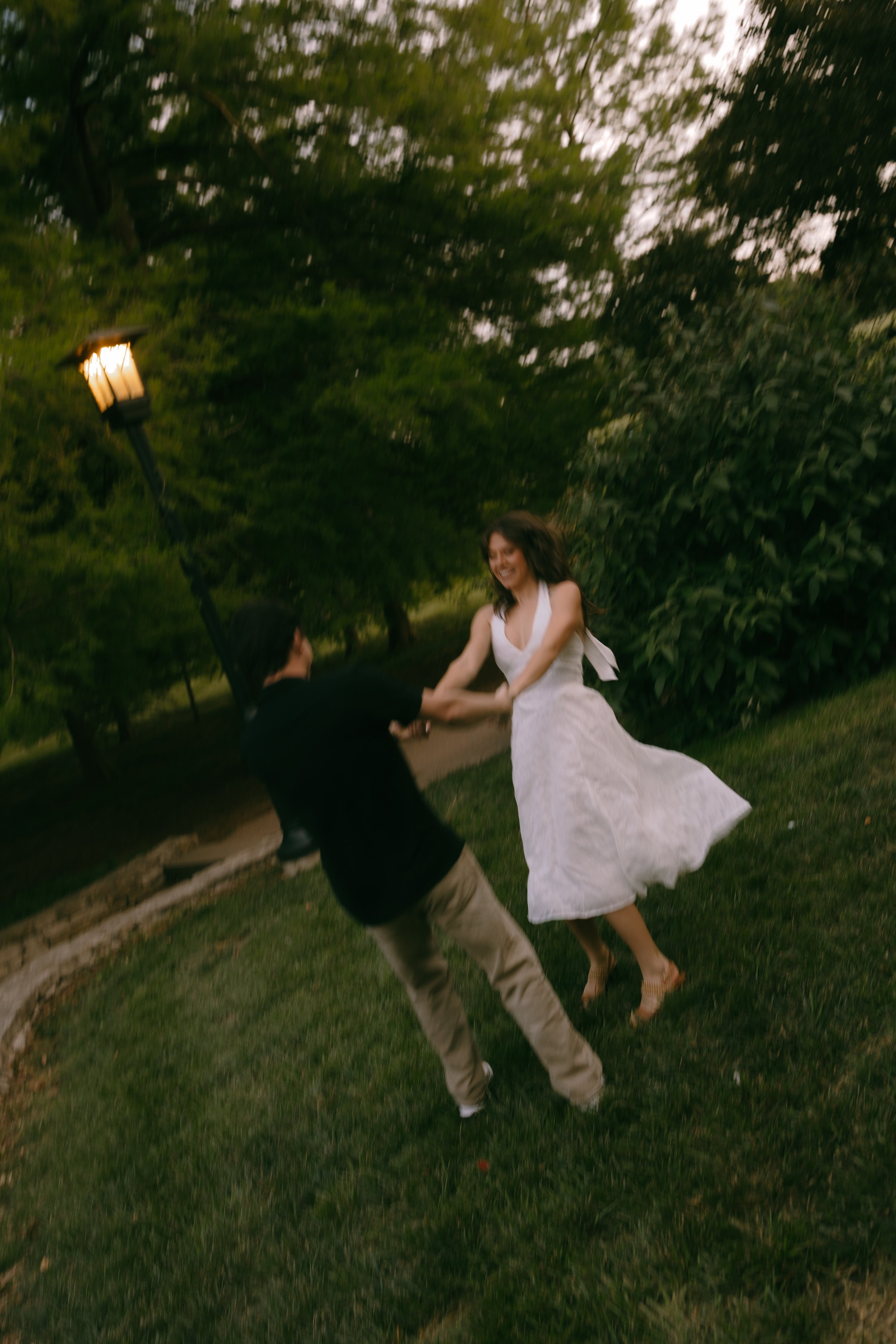 A couple has a playful dance in a garden, with the woman in a white dress spinning and smiling, and the man in dark clothing holding her hands, amidst trees and a lamppost.