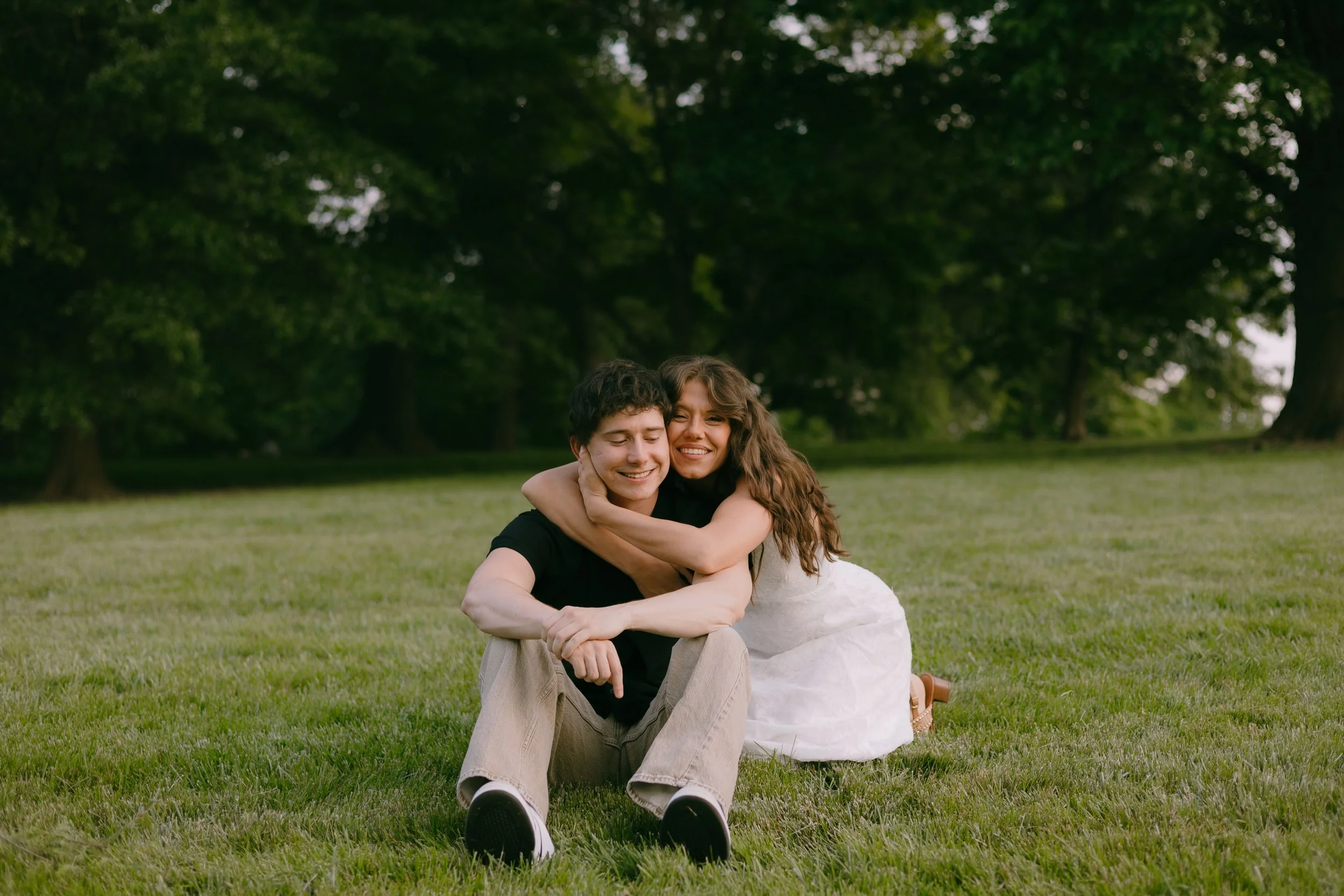 A young man and woman sitting on the grass in a park, hugging and smiling, with trees in the background.