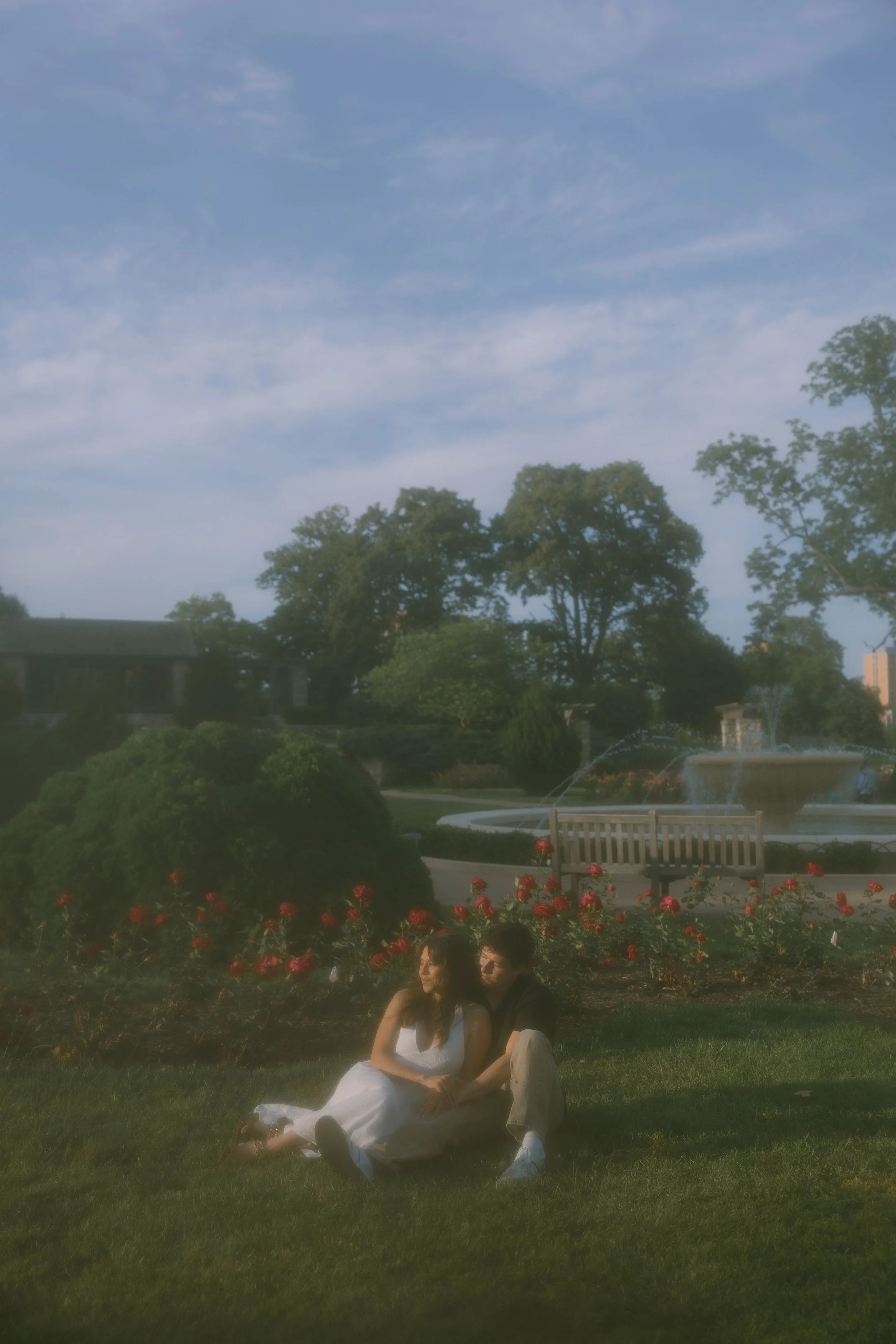A man and a woman sitting together on grass in a park, surrounded by red flowers with trees and a fountain in the background on a sunny day with a partly cloudy sky.