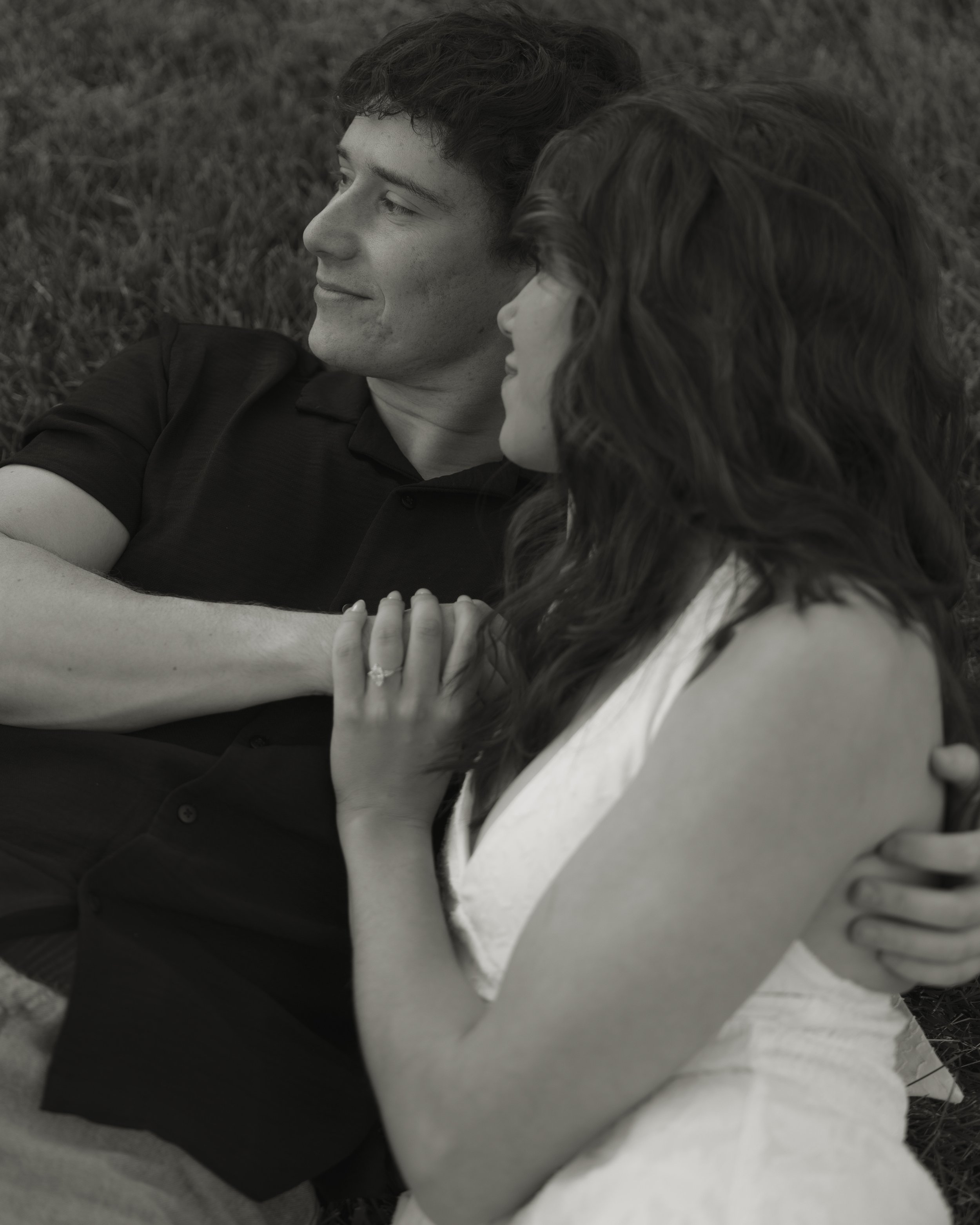 A young man and woman lie on the grass closely together, looking into each other's eyes with a smile, with their heads near each other.