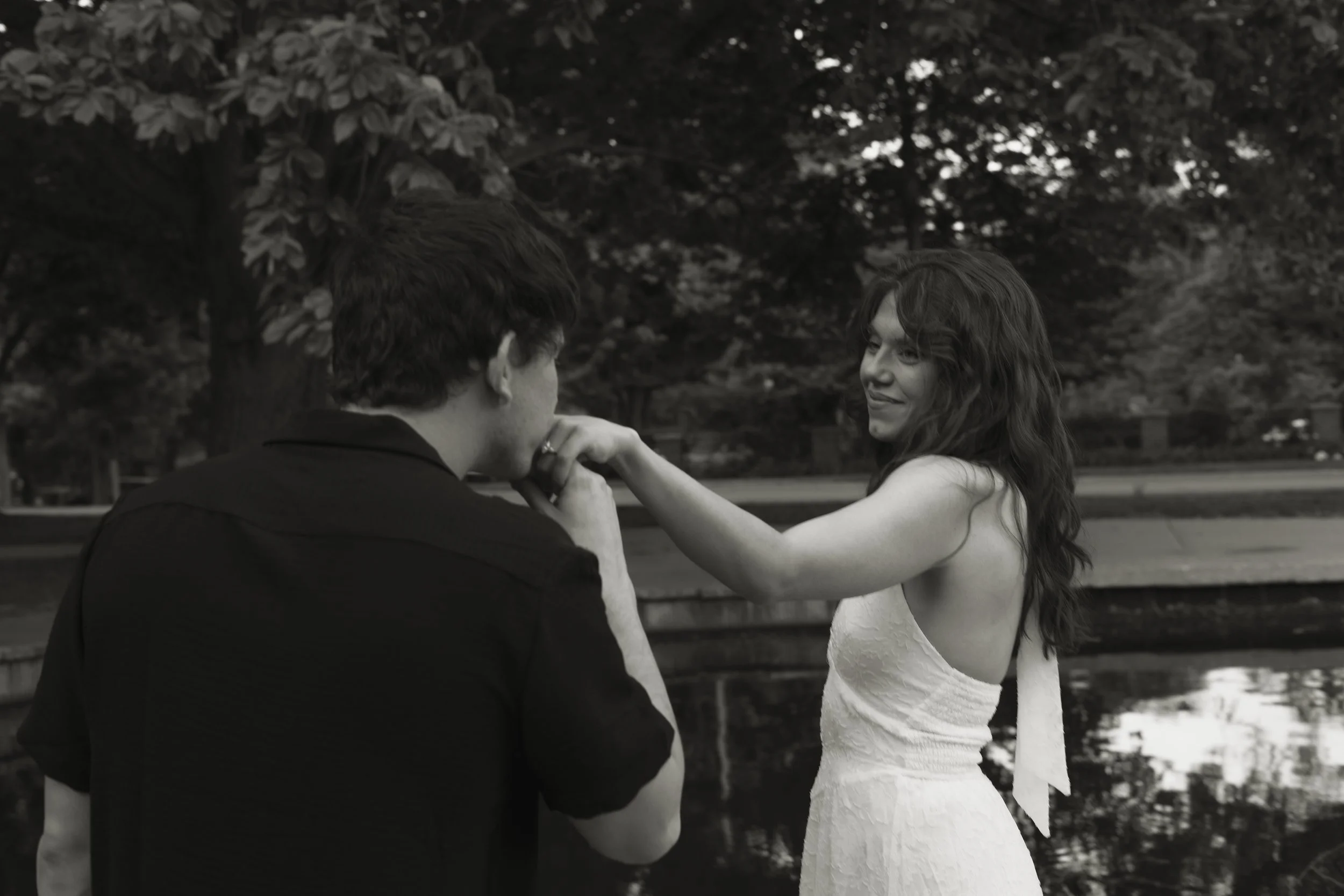 A black and white photo of a man and woman standing outdoors with trees in the background. The man is facing away from the camera, and the woman is smiling while looking at him, holding his hands.