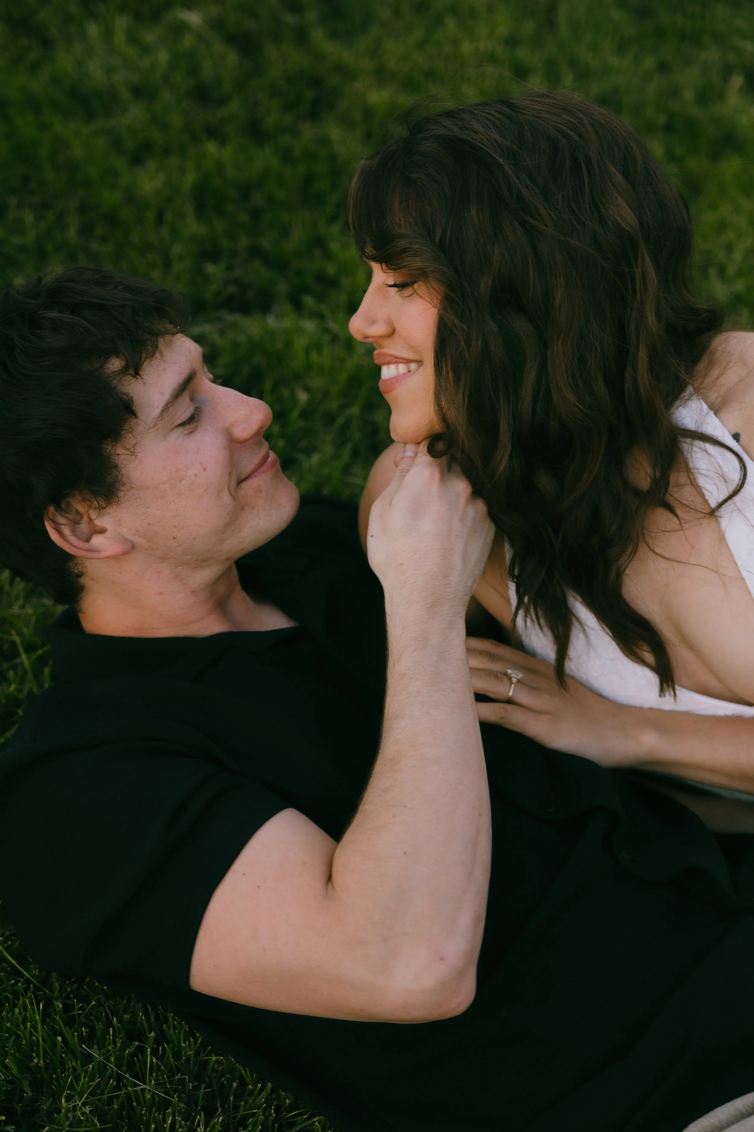 A young man and woman lying on green grass, the man looking up at the woman who is smiling at him.