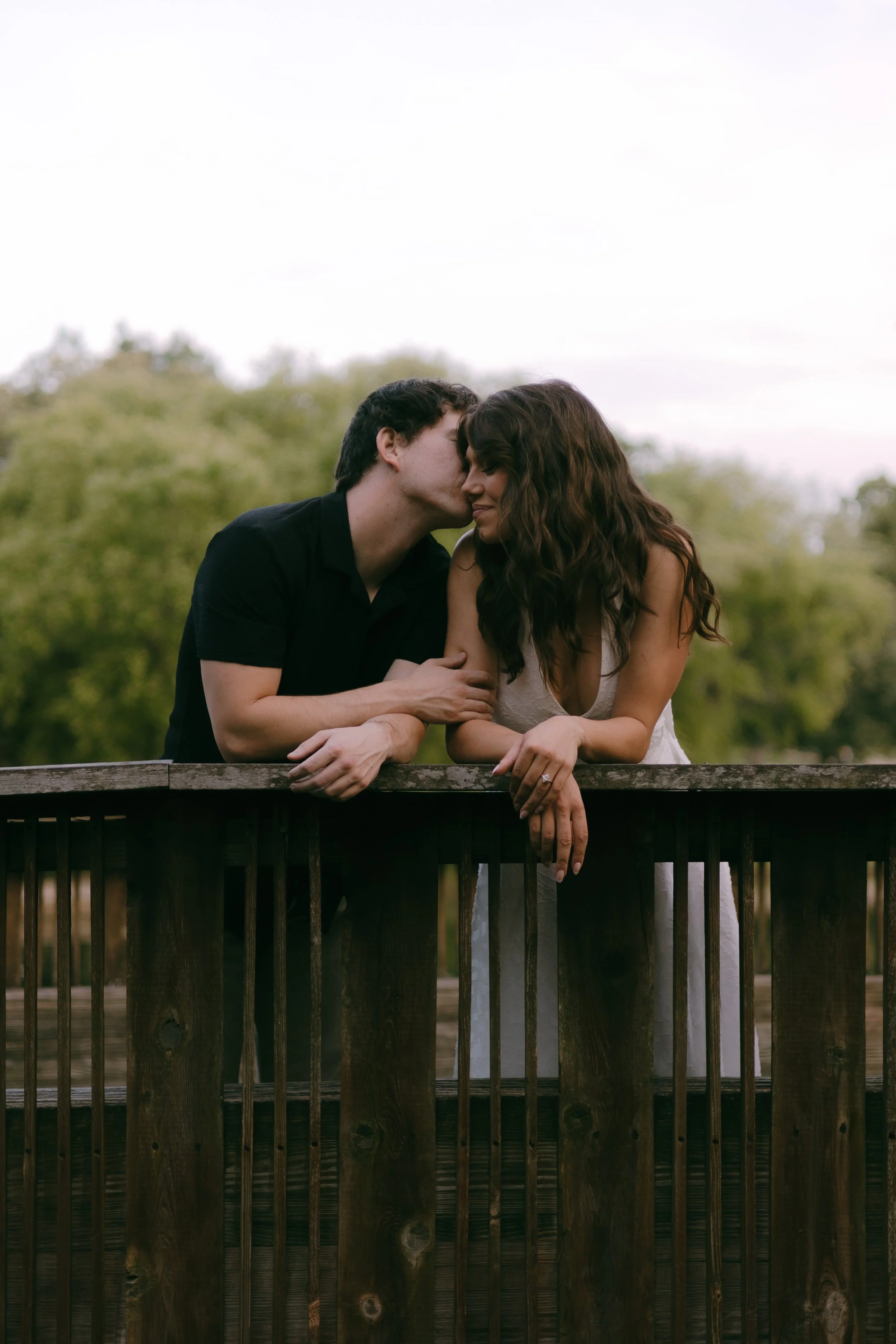 A young man and woman sharing an intimate moment outdoors, leaning into each other with their noses touching, standing behind a wooden fence with green trees in the background.