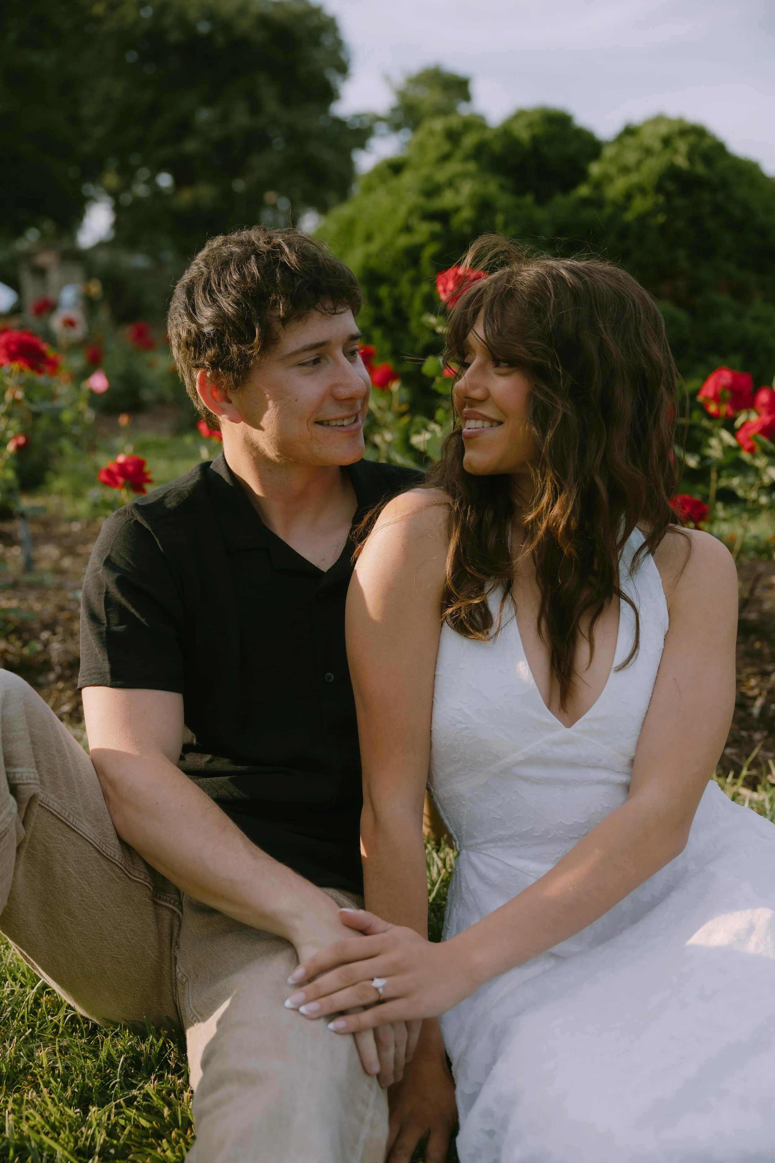 A couple sitting outdoors in a garden with red roses, holding hands and looking into each other's eyes, smiling.