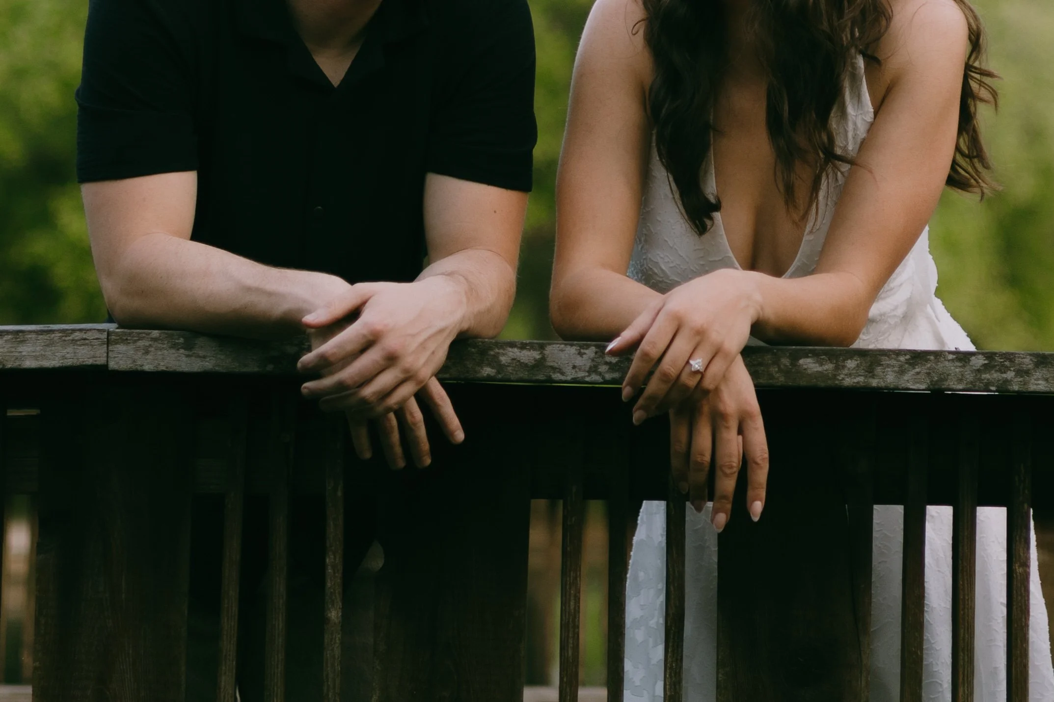 Close-up of two people standing behind a weathered wooden fence, their arms resting on top, with a blurred green outdoor background. The person on the left wears a black shirt, and the person on the right wears a sleeveless white dress and a ring on 