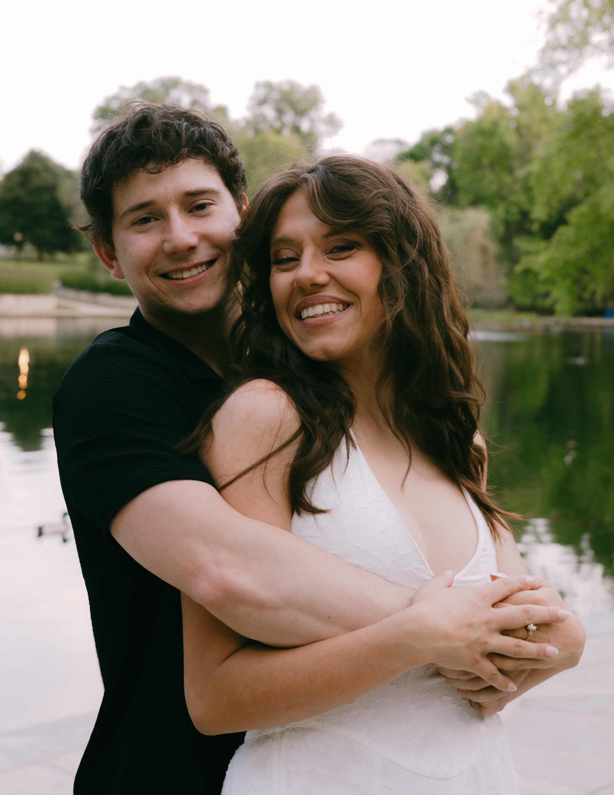 A smiling couple standing outdoors near a body of water, with trees in the background. The woman is in a white dress, and the man is in a black shirt, embracing each other.