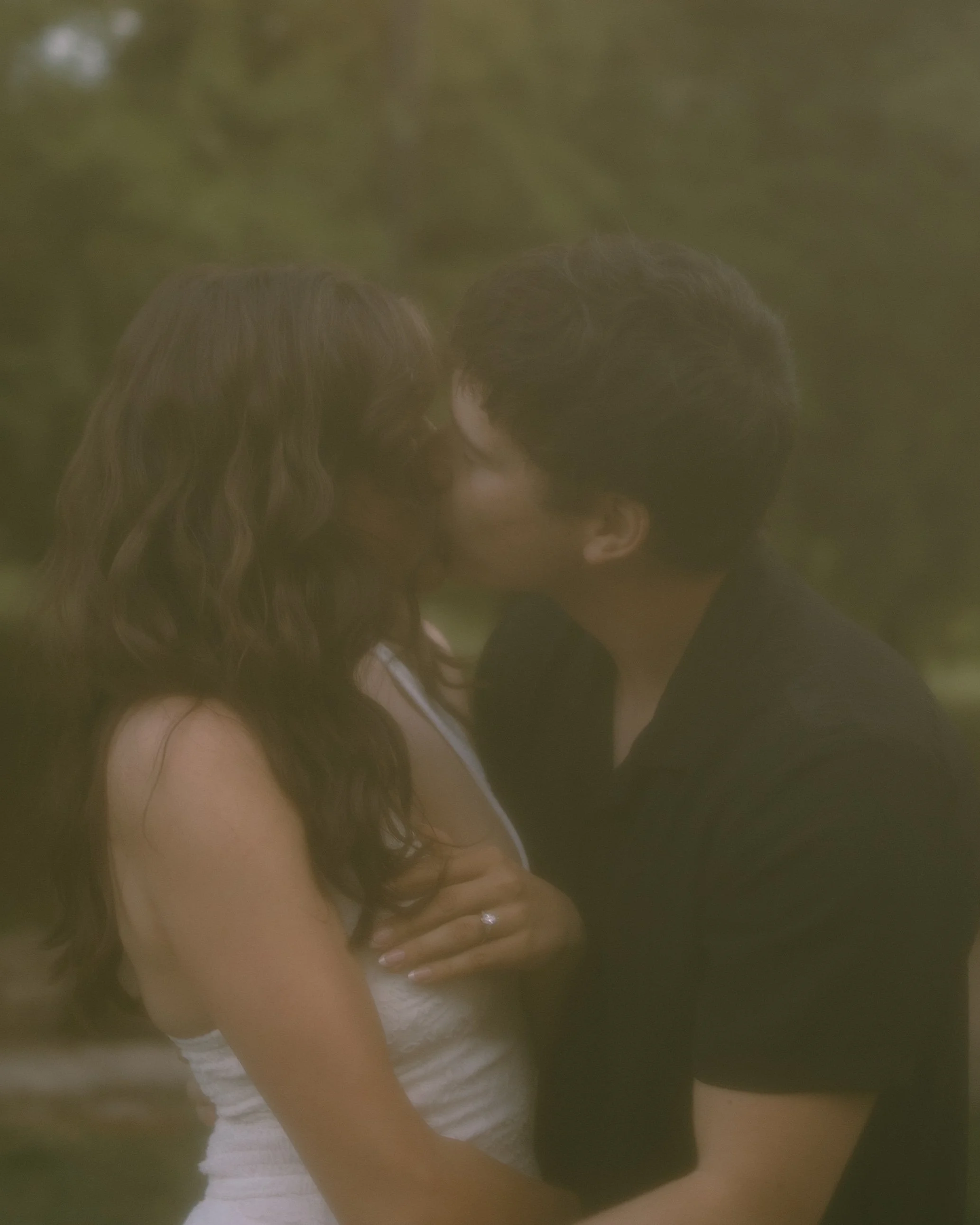 A woman with long wavy hair wearing a white tank top and a man with short hair and glasses wearing a dark shirt, are close together, possibly embracing or preparing to kiss, outdoors with greenery in the background.