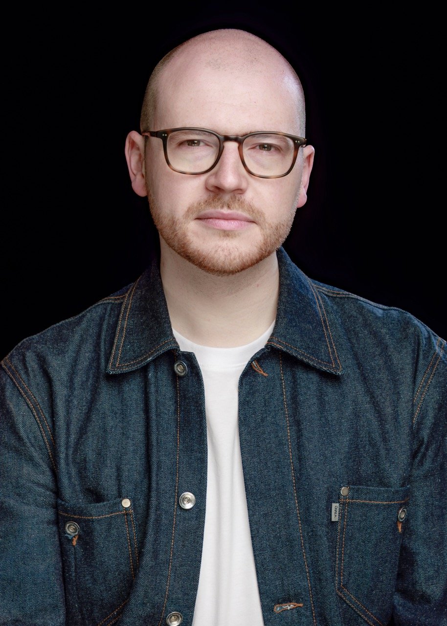 A portrait of a man with a shaved head, glasses, light facial hair, wearing a denim jacket and white shirt against a black background.