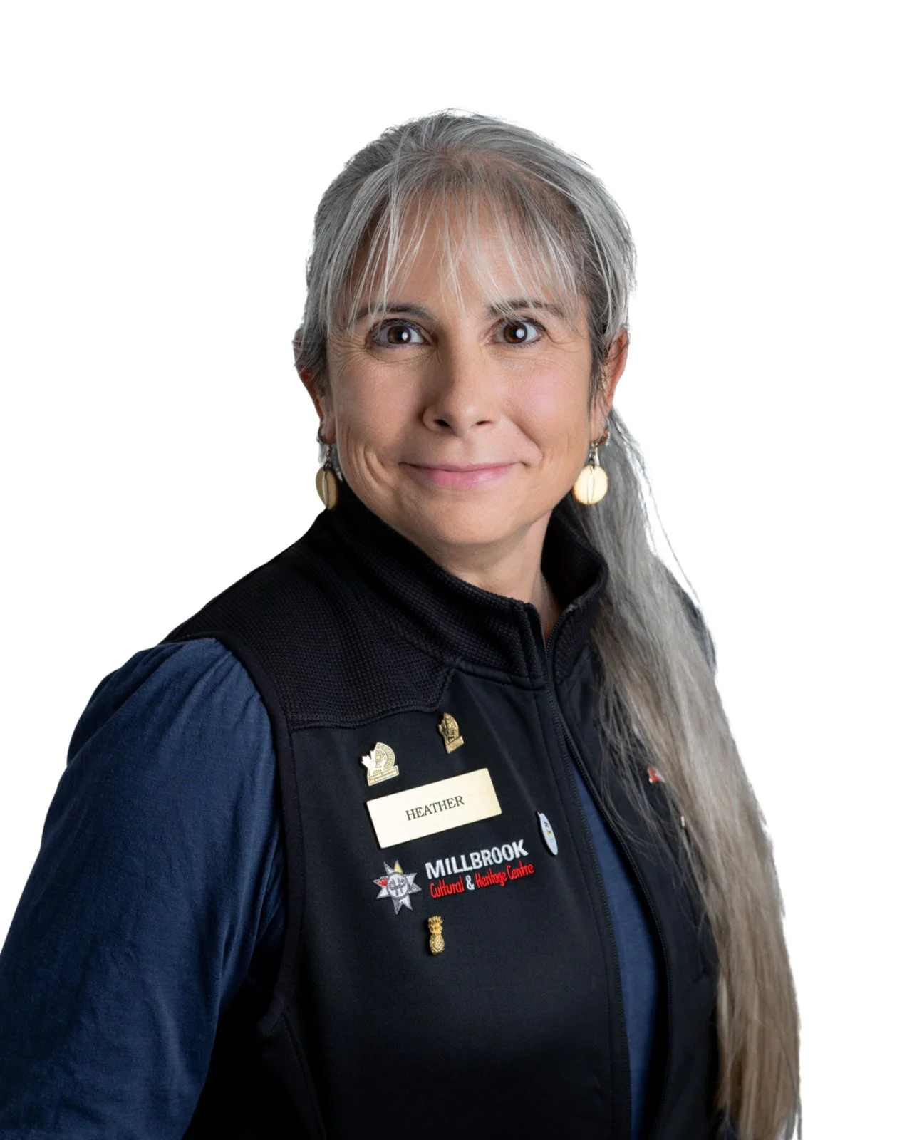 Portrait of a woman with long hair, wearing a black vest with badges and a name tag that says Heather, against a white background.