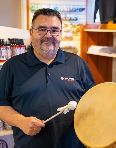 A man smiling while holding a mallet in a store, with shelves of bottles and products in the background.