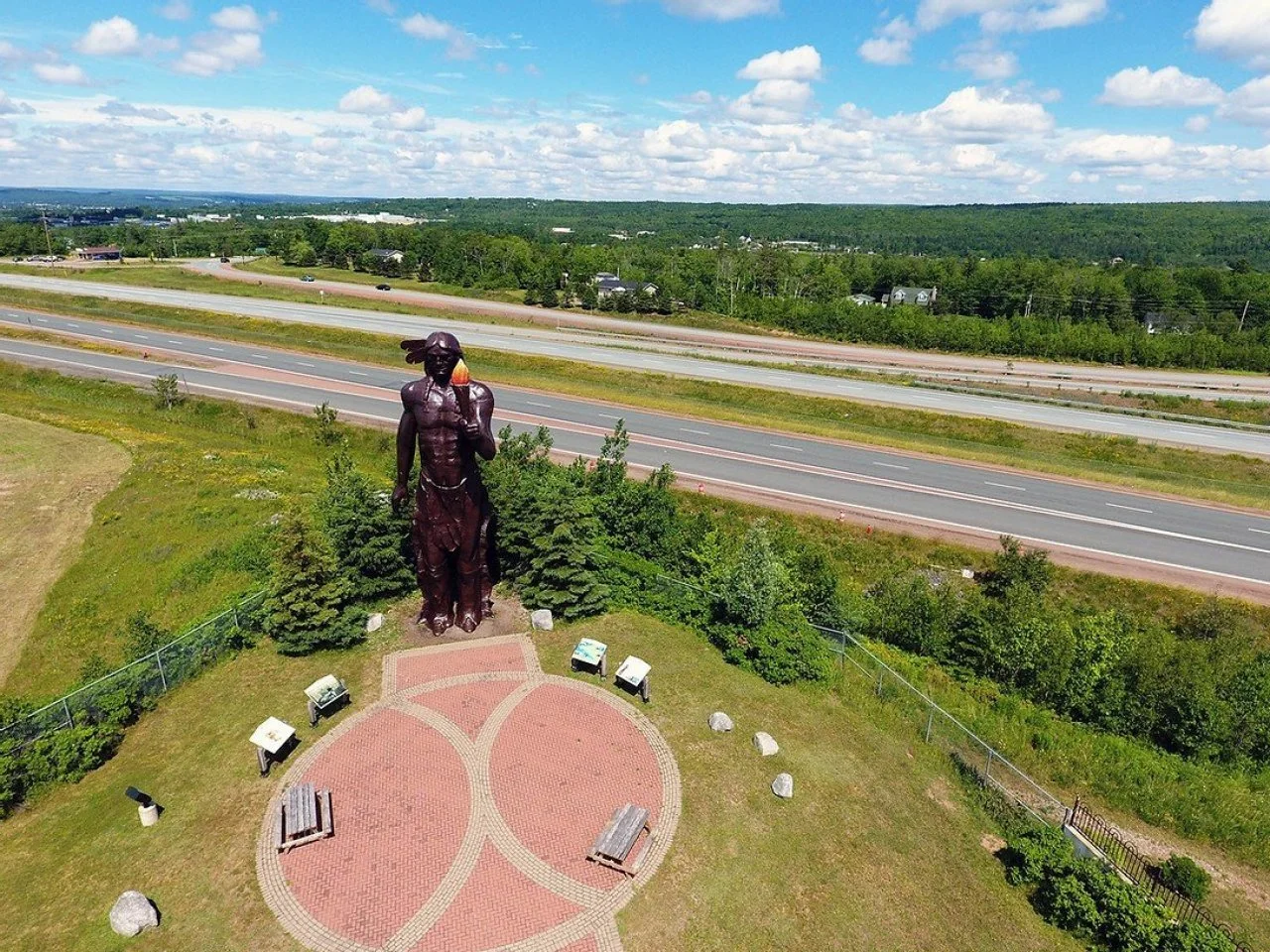 Large bronze statue of Glooscap, standing on a circular brick-paved area with benches and trees, near a highway with green landscape and a cloudy sky.