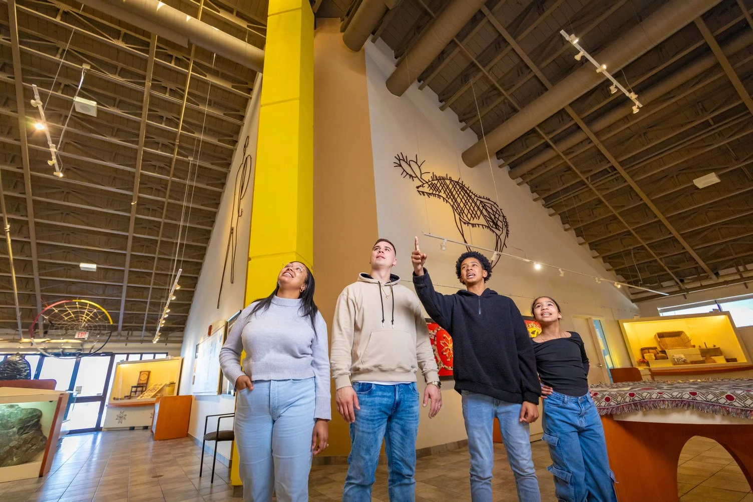 Four young people standing inside a museum, looking upward, with one person pointing. The museum has artwork on the walls, including a large outline of a moose and other displays.