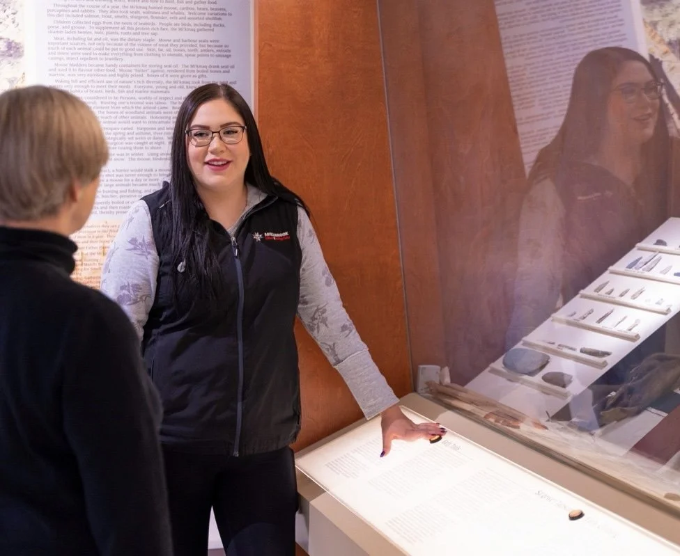 A woman with long dark hair, glasses, and a black vest talking to a man with blond hair at a museum exhibit. She is standing beside a display case with artifacts and information panels.
