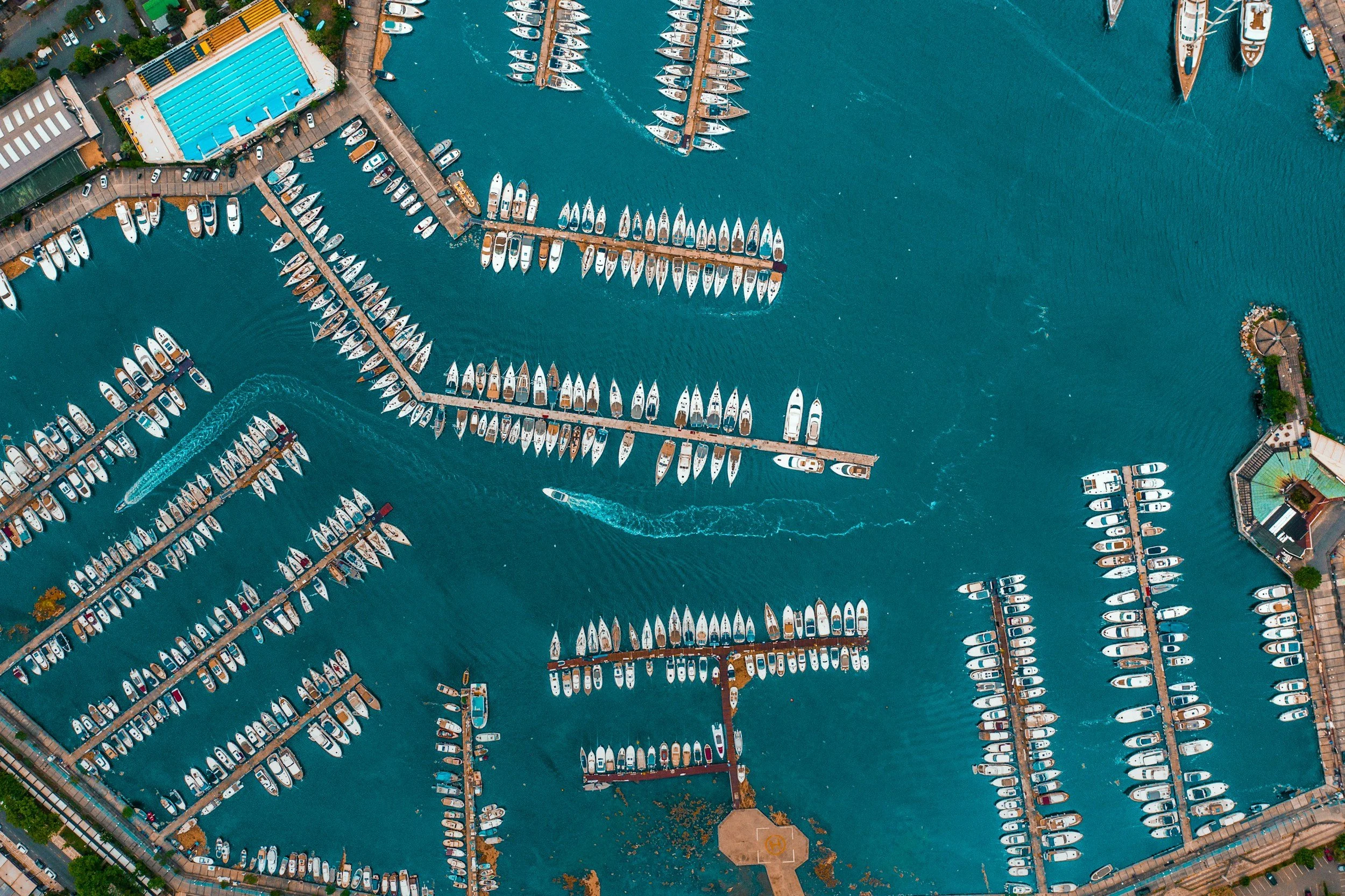Aerial view of a marina with numerous boats docked along the piers and a large swimming pool nearby, surrounded by buildings and green spaces.