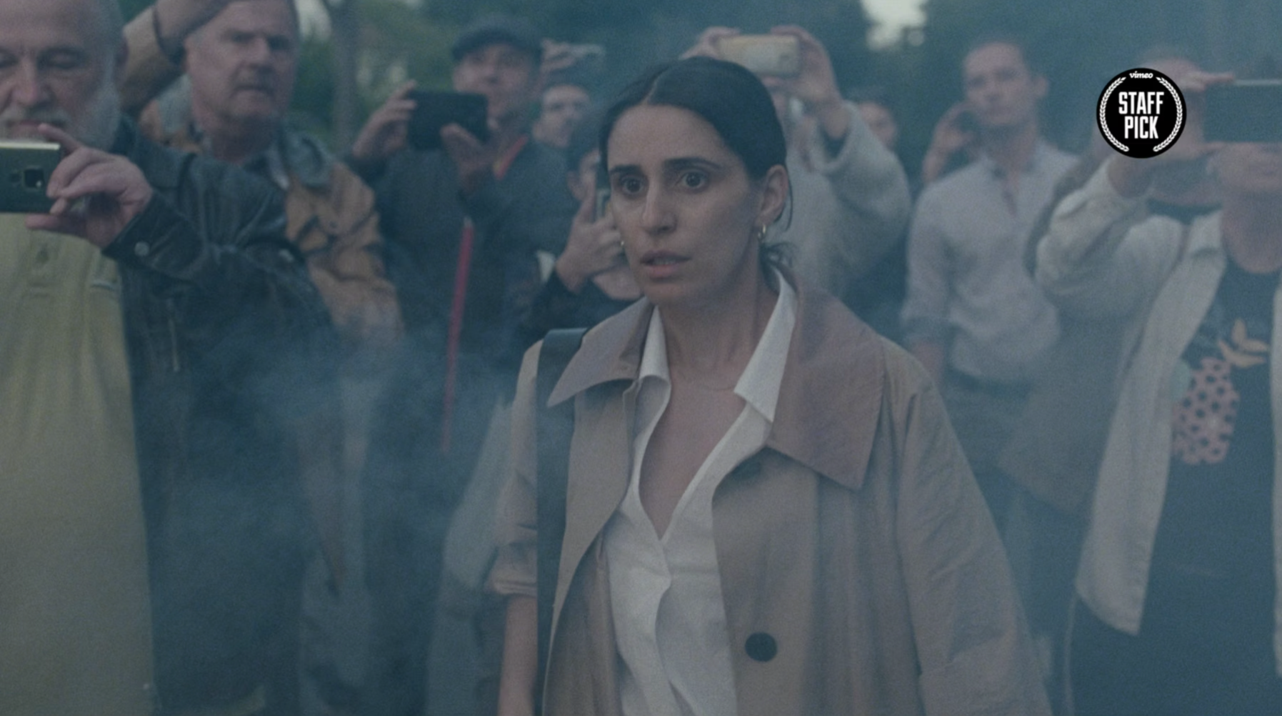 Eine Frau in einem beige Trenchcoat steht vor einer Gruppe von Menschen, die Fotos machen, während sie durch Rauch oder Nebel schaut.