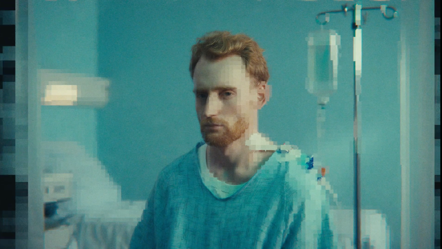 Ein Mann mit rotem Haar und Bart trägt einen Krankenhaus-Grotxe und steht vor einem Krankenhausschrank, das Bild ist digital stark verpixelt.