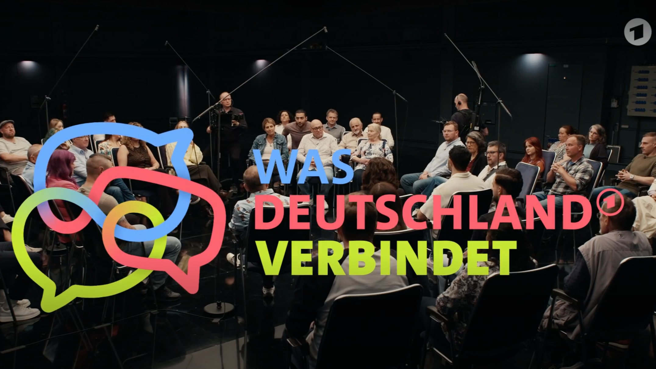 Mehrere Menschen sitzen in einem dunklen Raum während einer Veranstaltung. Über dem Bild sind bunte Sprechblasen und der deutsche Text 'WAS DEUTSCHLAND VERBINDET' zu sehen.