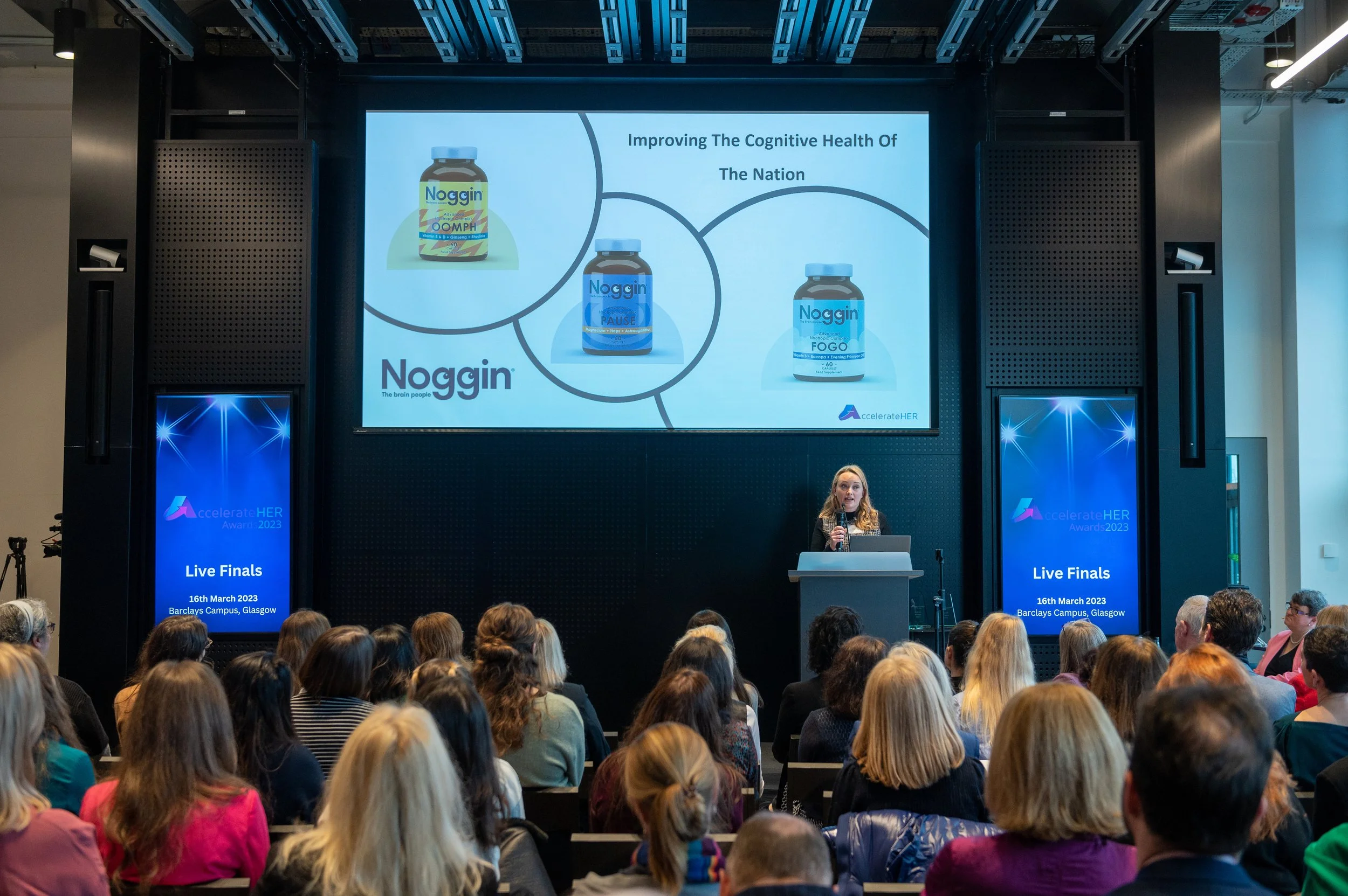A presentation slide about improving the cognitive health of the nation, showing three bottles of Noggin supplements and a speaker standing at a podium in front of a large audience at Barclays Campus in Glasgow during the 16th March 2023 event.