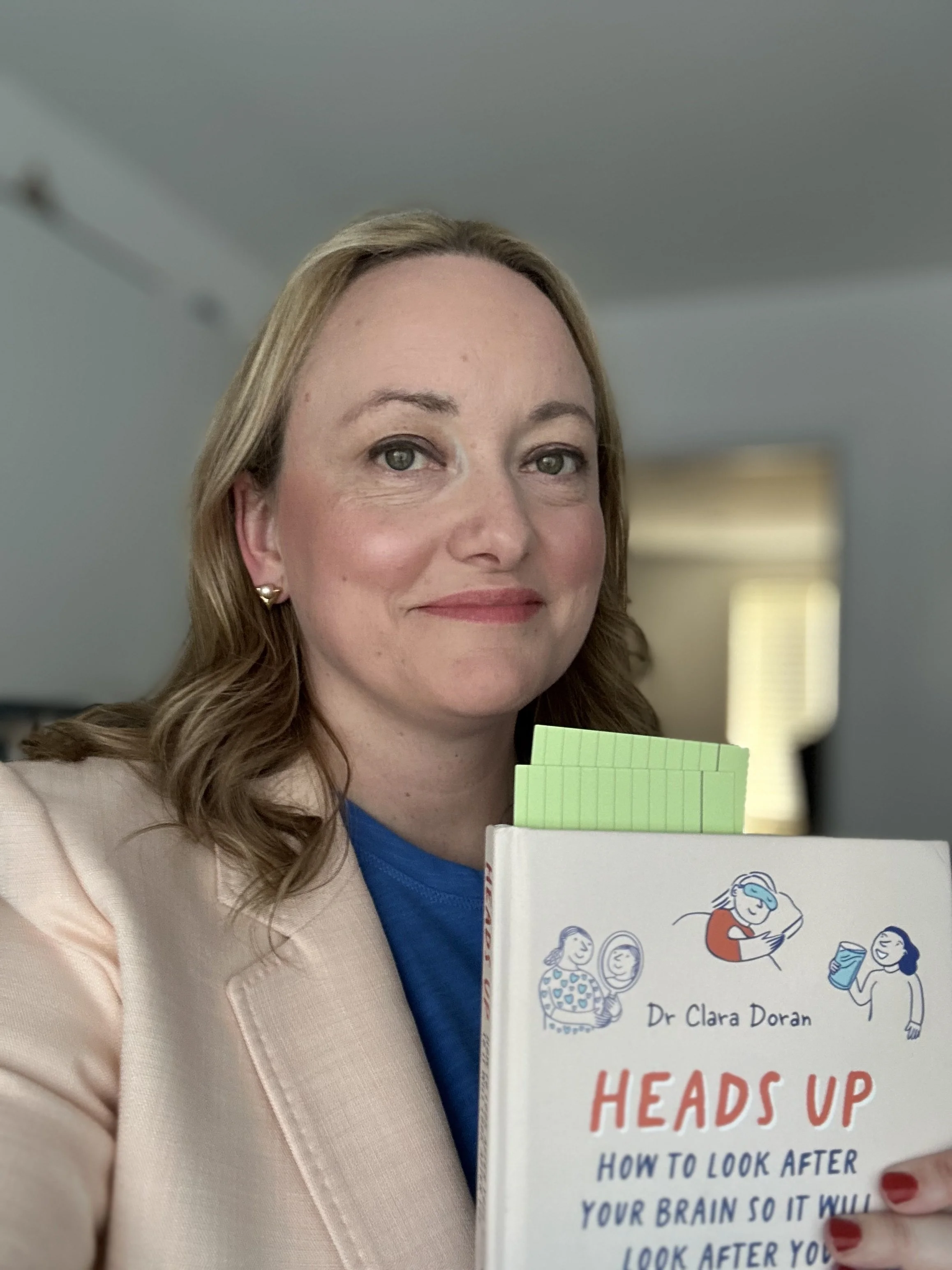 A woman with wavy brown hair, wearing a beige blazer and blue top, is holding a book titled 'Heads Up' by Dr. Clara Doran. The book cover features cartoon illustrations of women and text about how to improve brain health.