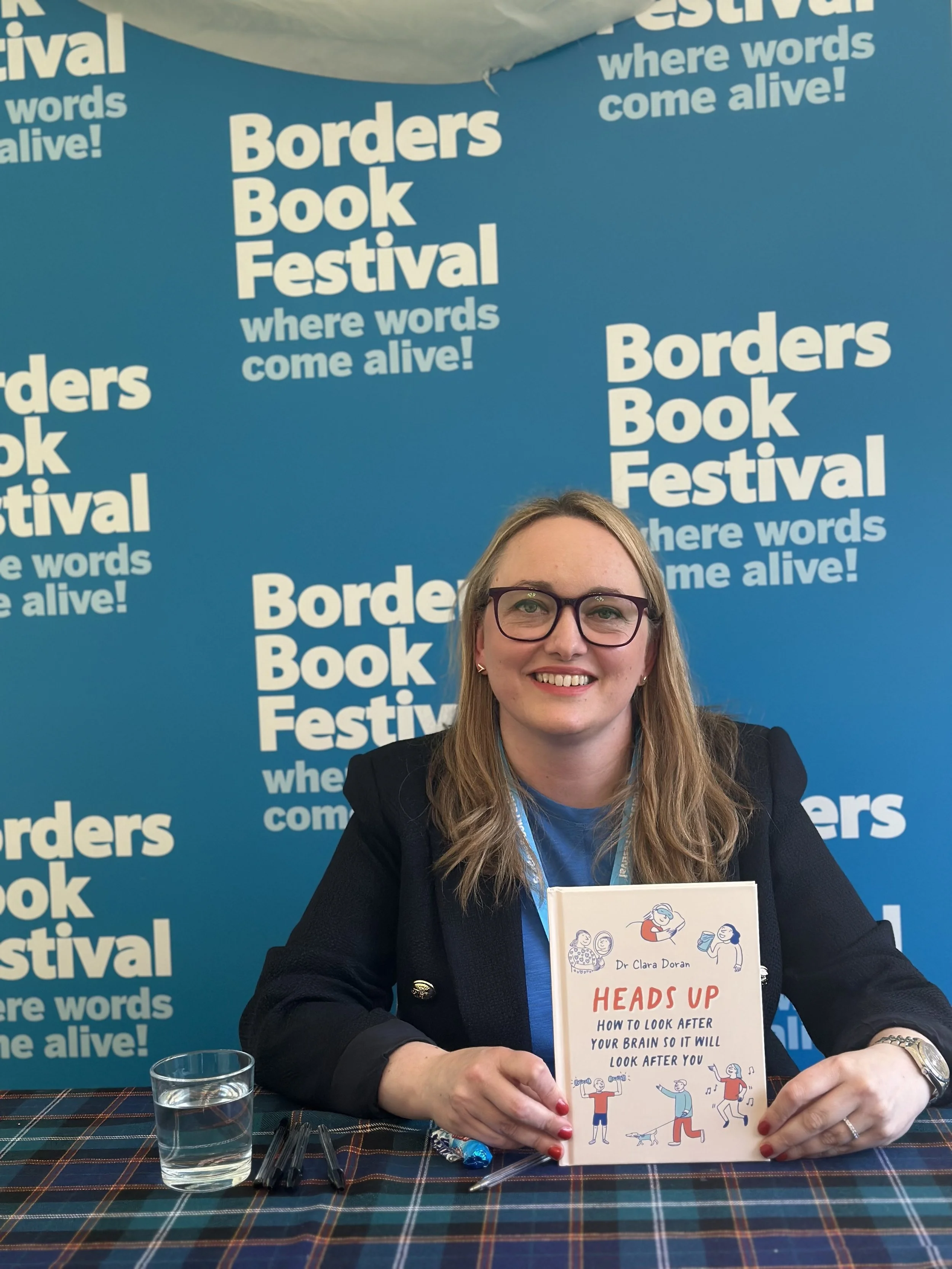 A woman with blonde hair, glasses, and a black jacket, smiling and holding an illustrated book titled 'HEADS UP' at a book festival. The backdrop is a blue wall with repeating white text promoting the Borders Book Festival and its tagline.