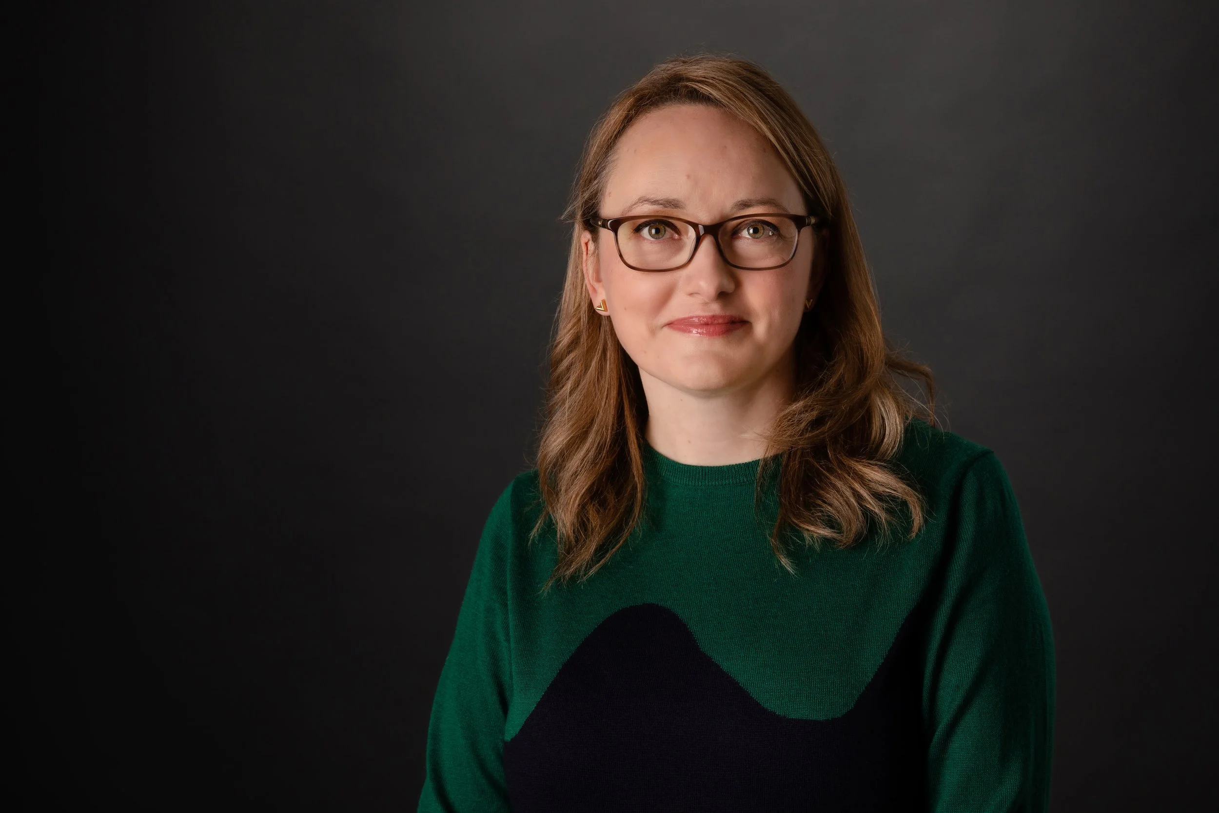 Portrait of a woman with brown hair, glasses, and a green sweater against a dark background.