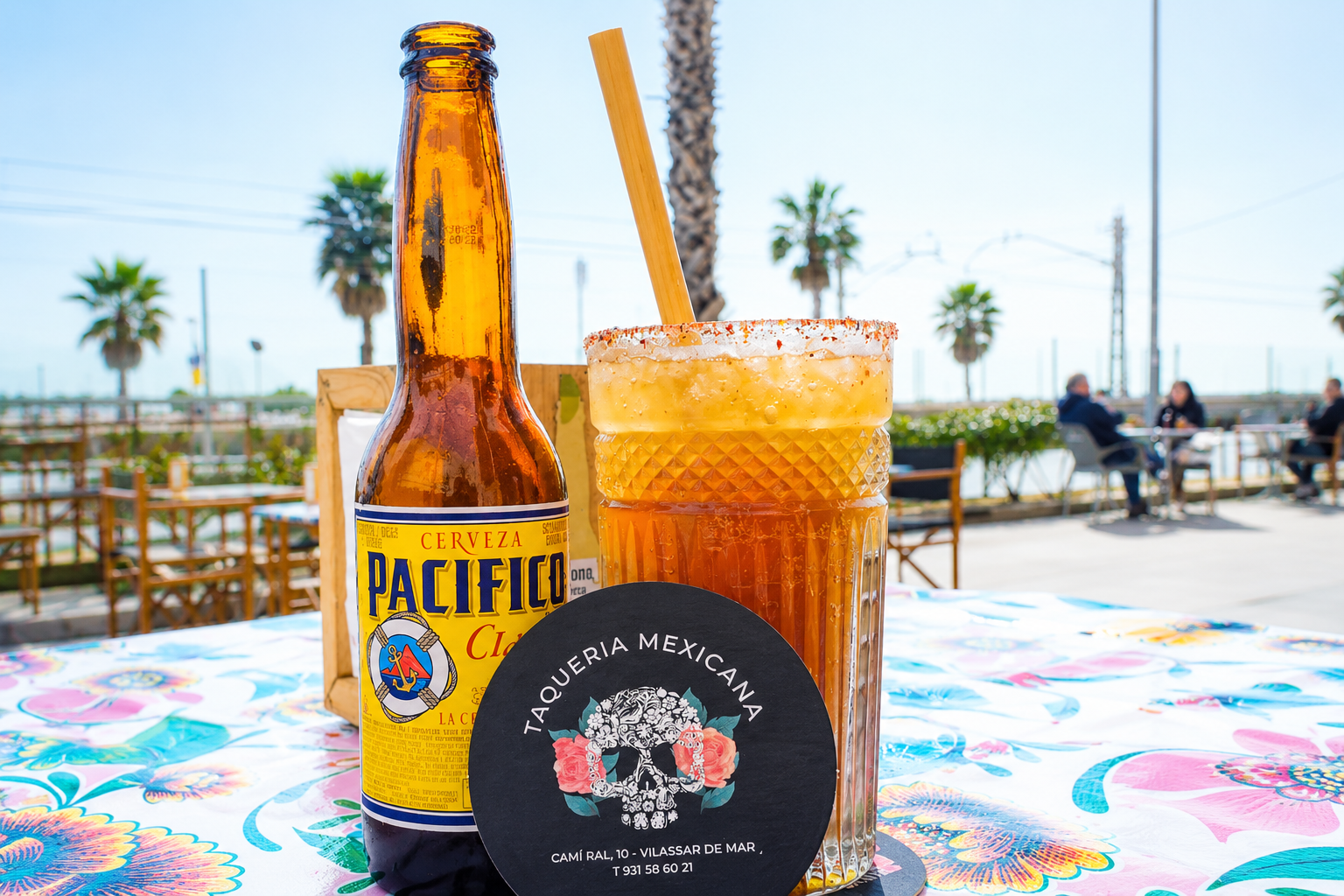 Una botella de cerveza Pacifico, un vaso con una bebida de color naranja con sal en el borde y una pajilla de madera, en una mesa con mantel colorido, en un lugar con palmeras y personas en el fondo.