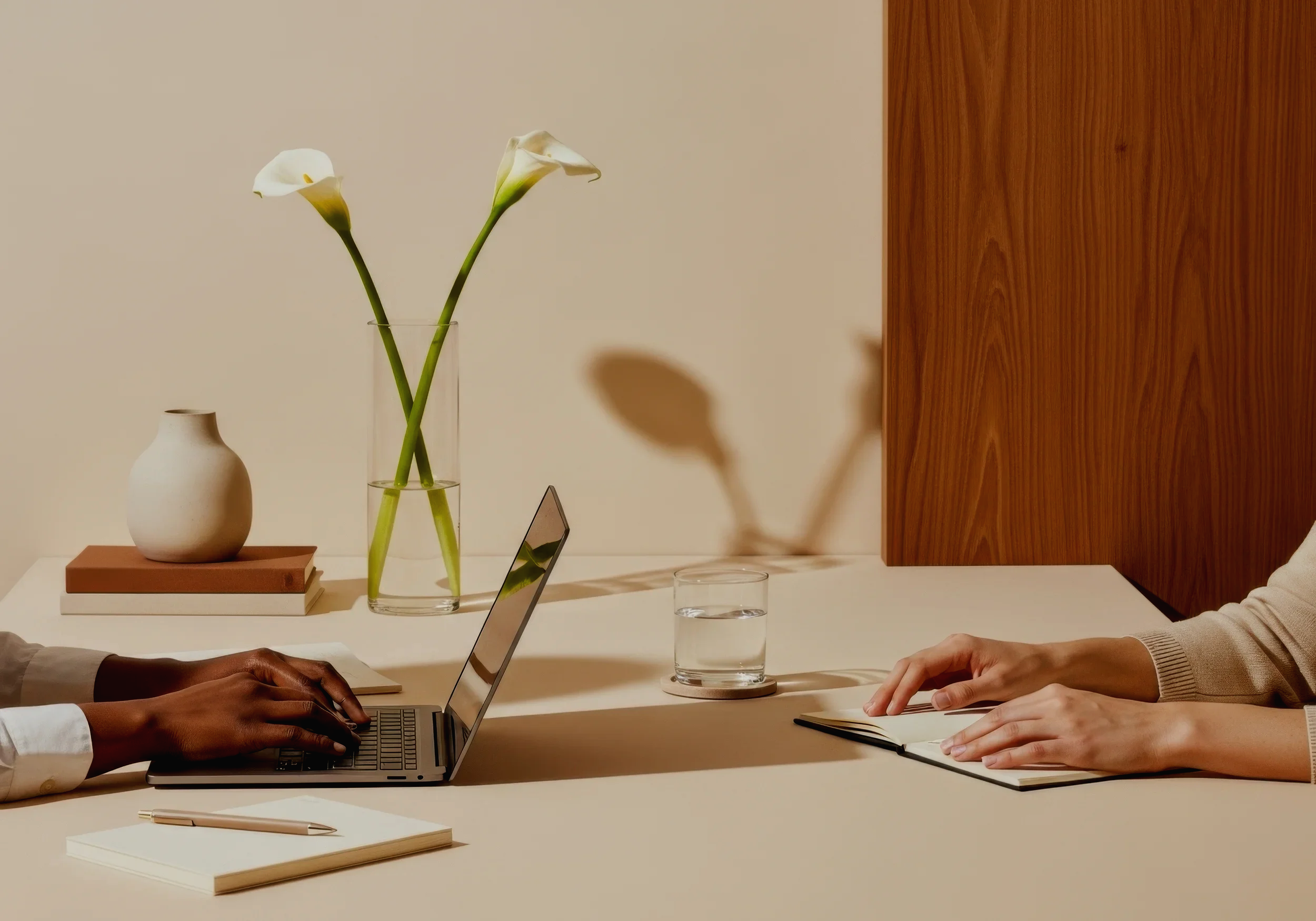 Two people sitting at a table working on laptops with notebooks and glasses of water, a wooden panel wall, and a vase with white calla lilies on the table.
