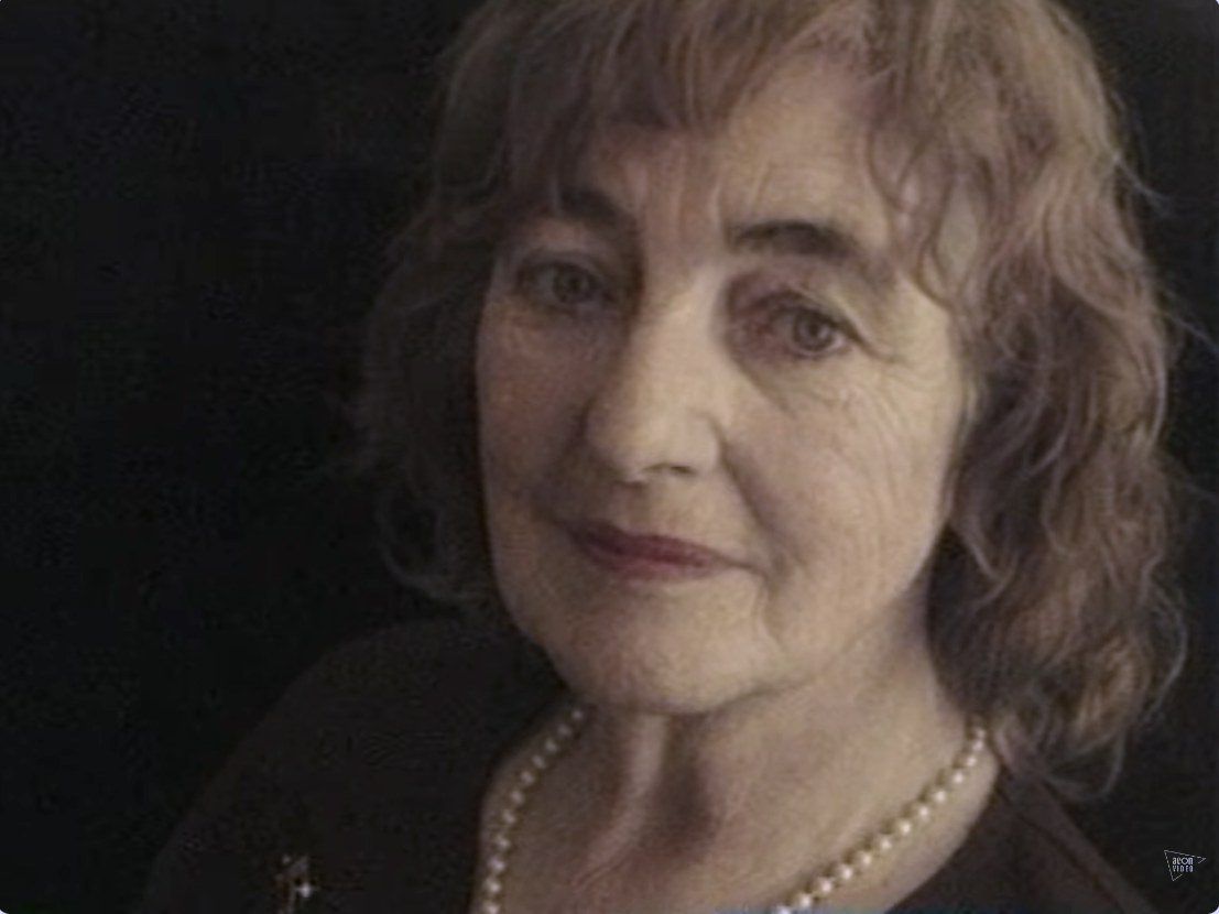 A portrait of an elderly woman with curly brown hair, wearing a black top and a pearl necklace, smiling softly against a dark background.