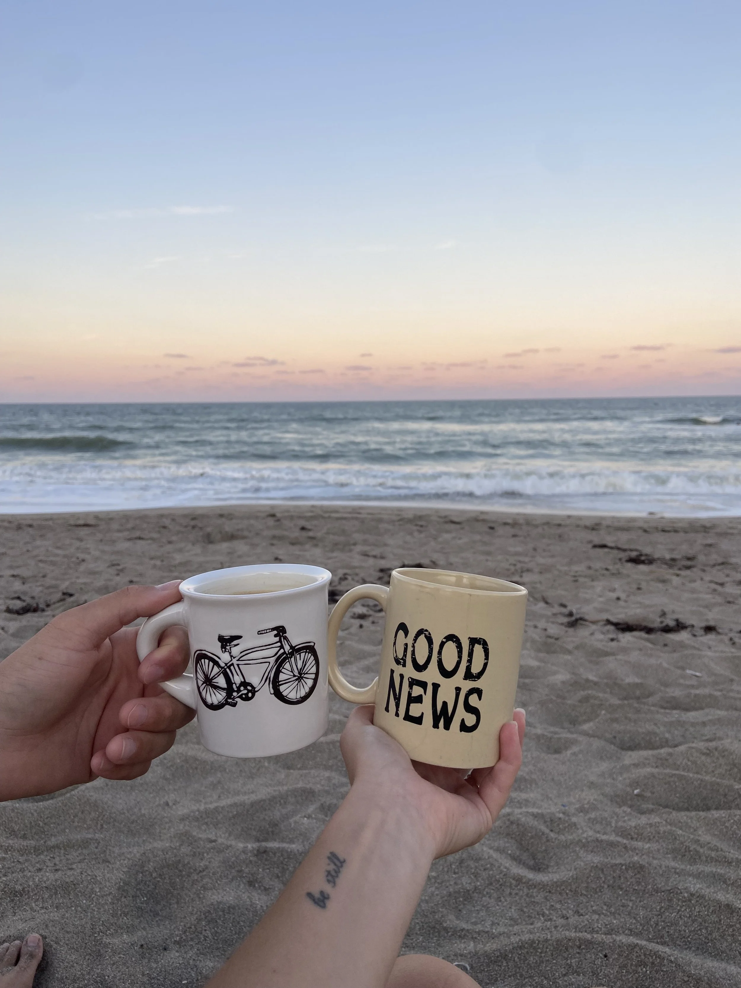 Two hands holding coffee mugs with a beach and ocean in the background, at sunset or sunrise.