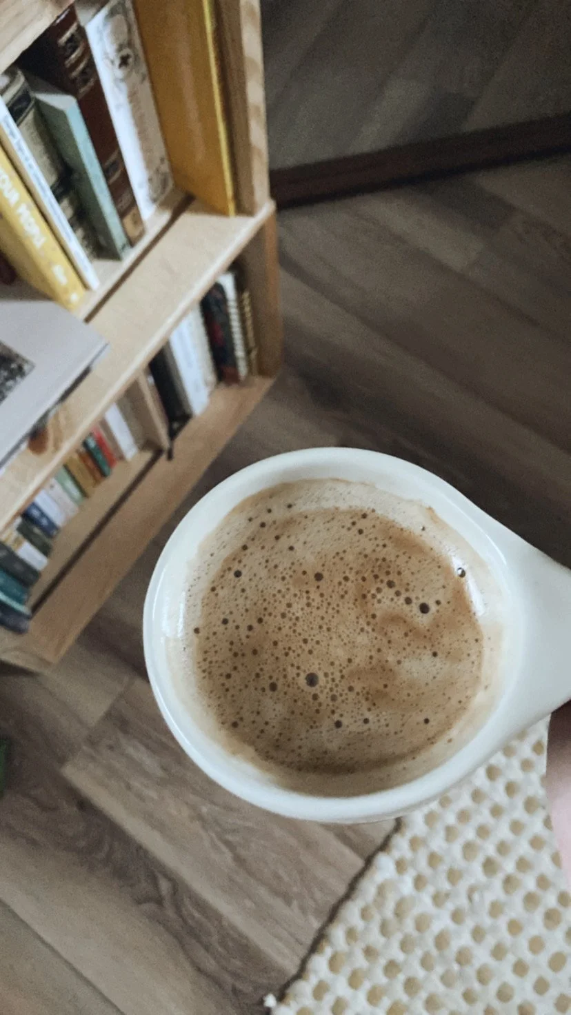 A cup of coffee with foam on top, held above a wooden surface in a room with a bookshelf in the background.