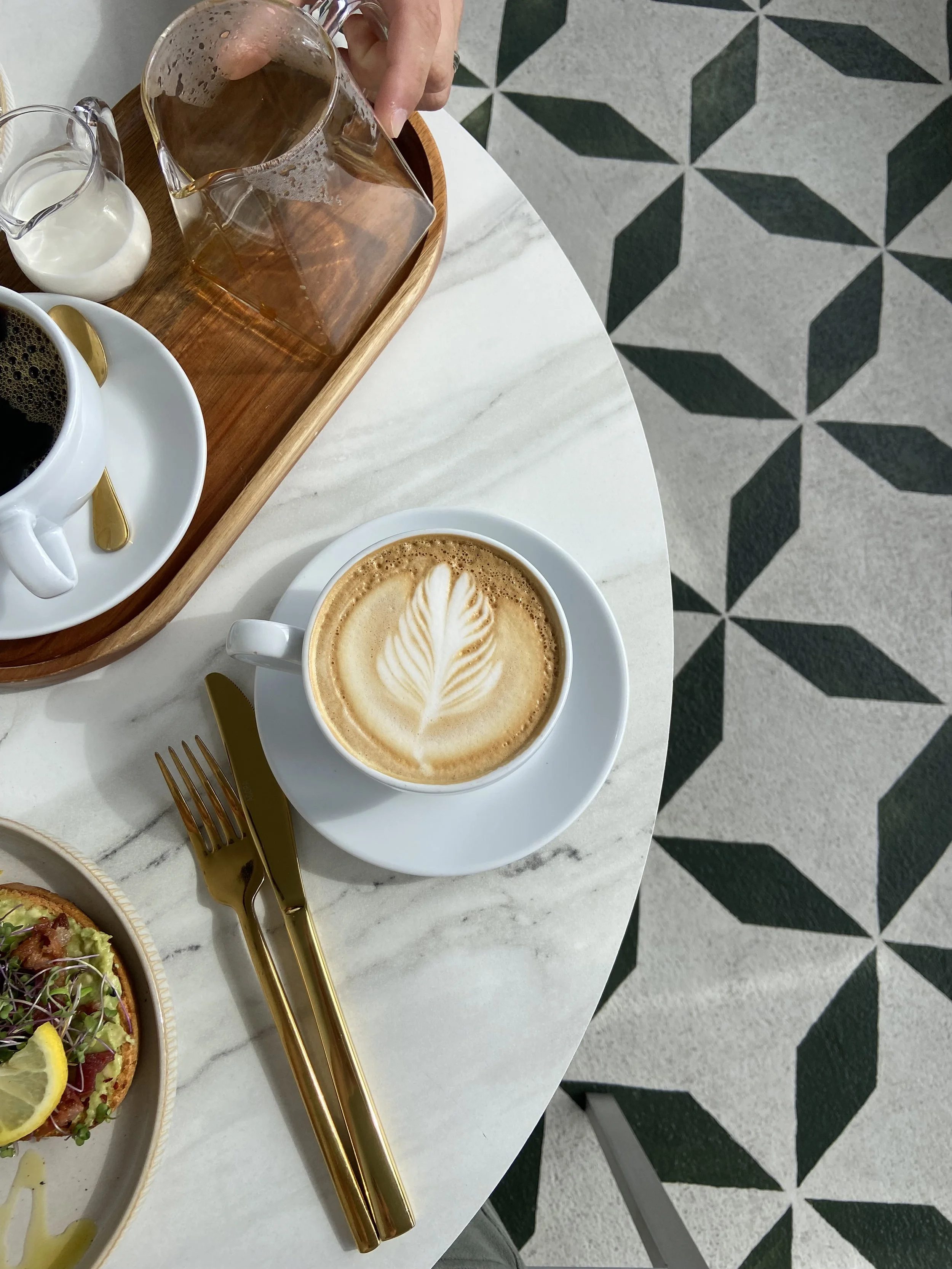A white marble table with a cup of cappuccino with latte art, a glass of iced tea, a small pitcher of cream or milk, and some gold-colored cutlery, with a patterned black and white tile floor in the background.