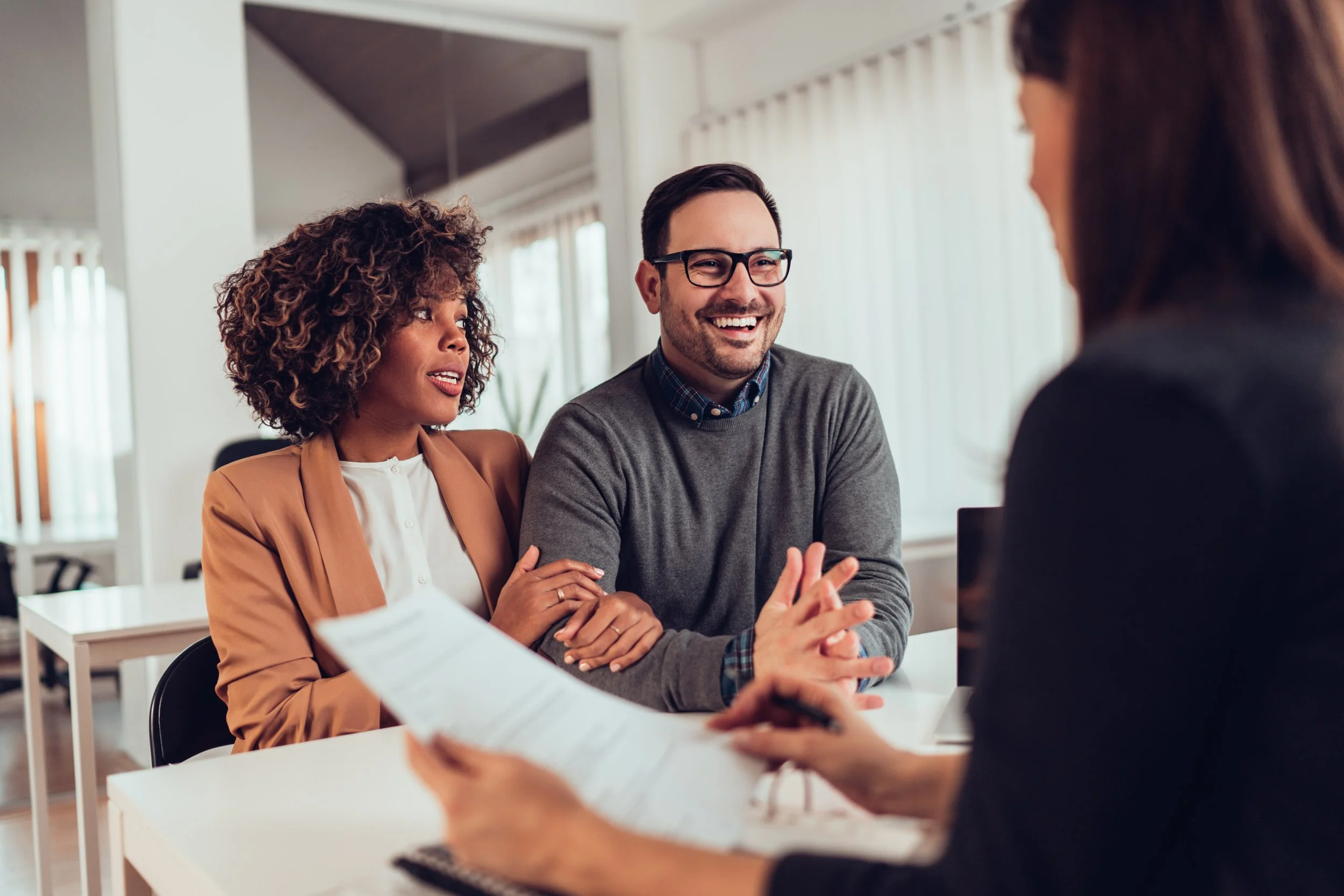 Three people having a discussion in a bright, modern office. One woman with curly hair and a tan blazer, a man with glasses and a grey sweater, and a woman with long brown hair are engaged in conversation around a white table with documents.