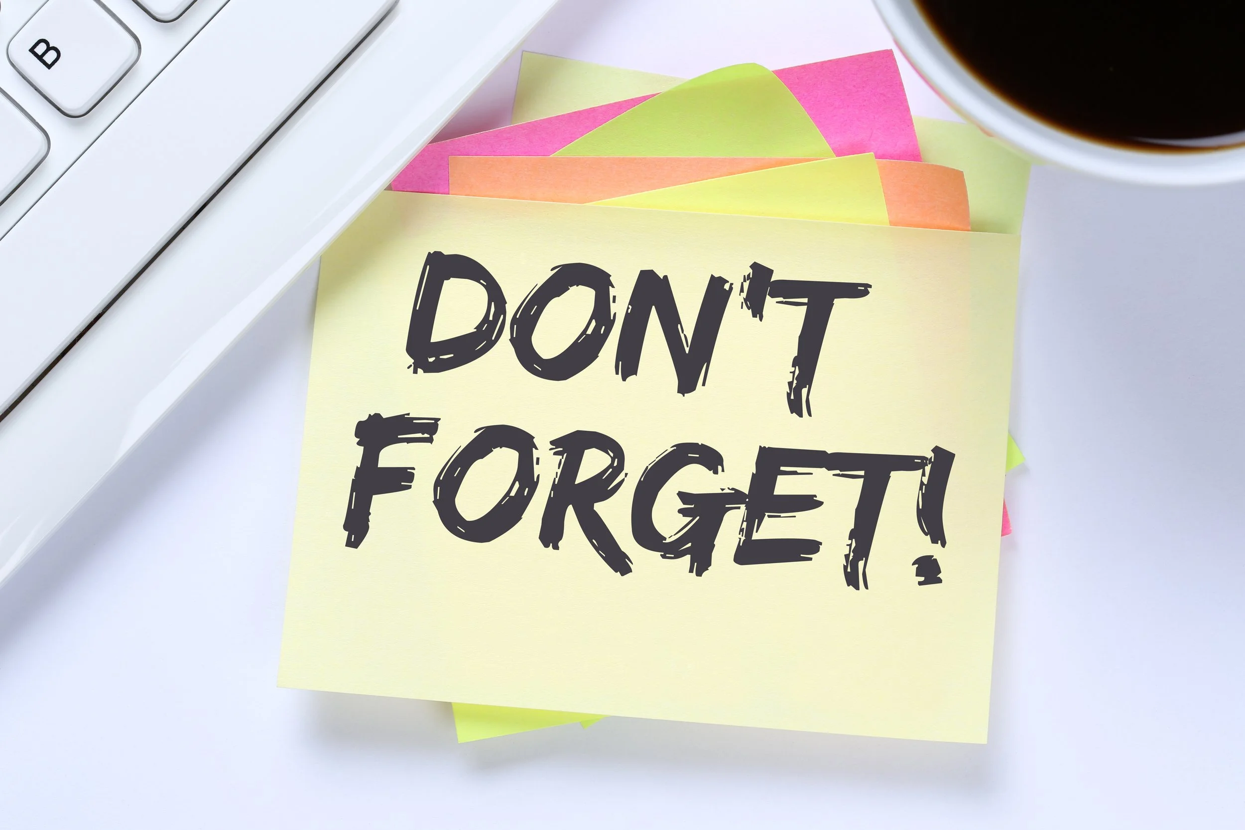 A computer keyboard partially visible on the left side of the image. In the center, there are sticky notes in yellow, pink, orange, and green with the words 'DON'T FORGET!' written on a yellow sticky note in bold black letters. In the top right corner, part of a cup filled with black coffee is visible.