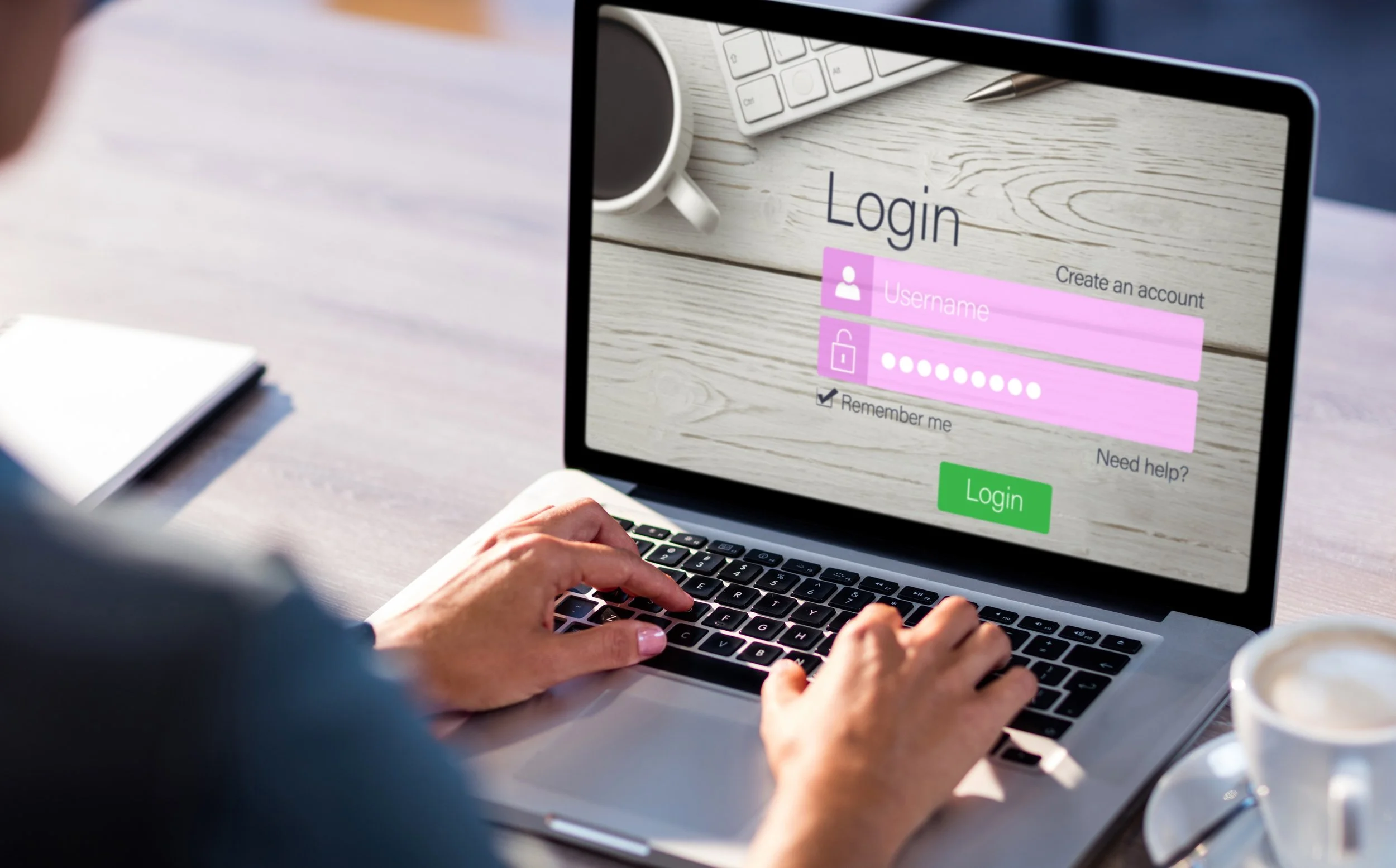 Person using a laptop to log into an account, with a login screen displaying fields for username and password, on a desk with a white notebook, a white mug, a keyboard, and a pen.