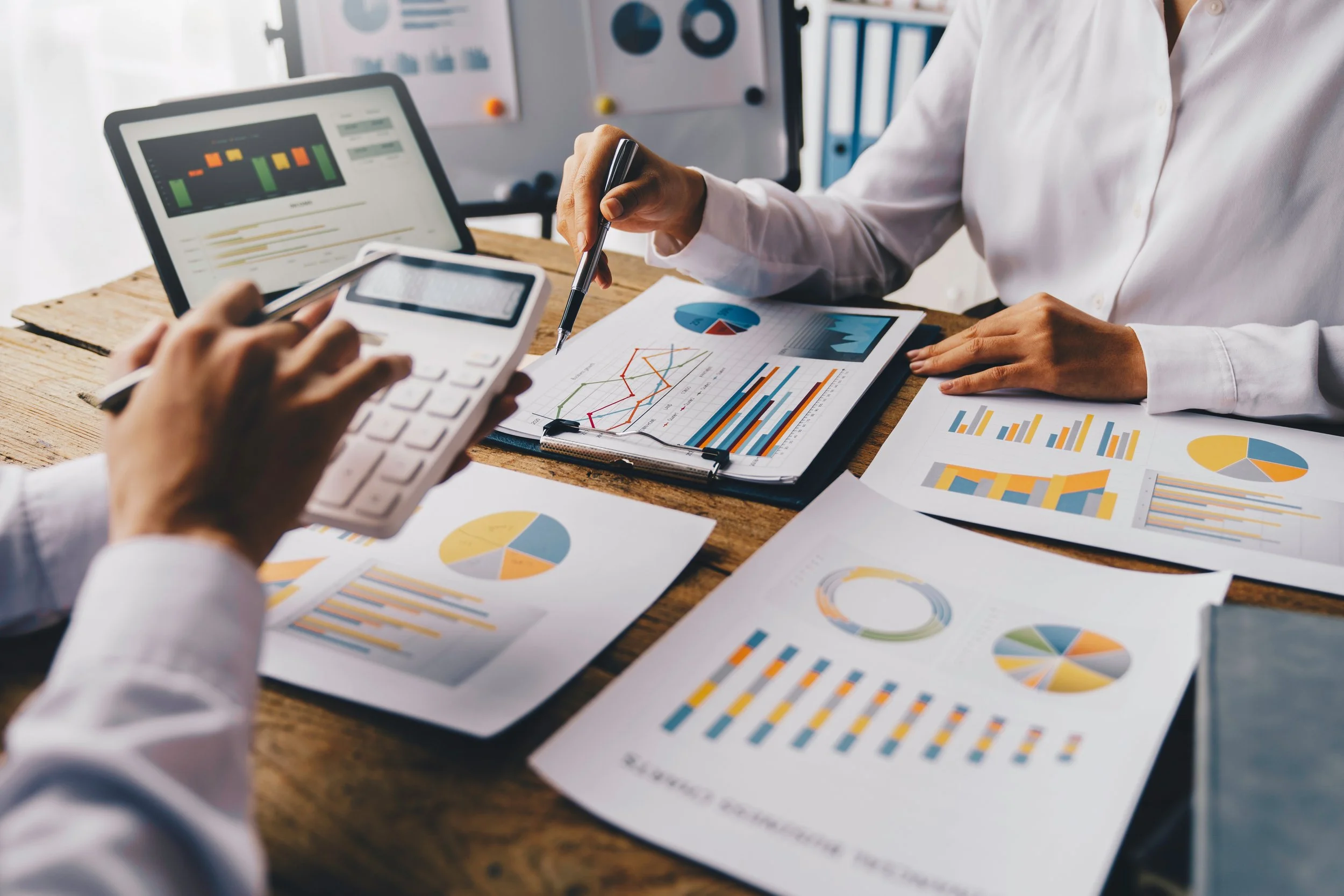 People working on a business meeting or project, analyzing various charts and graphs with financial data on papers and electronic devices on a wooden table.