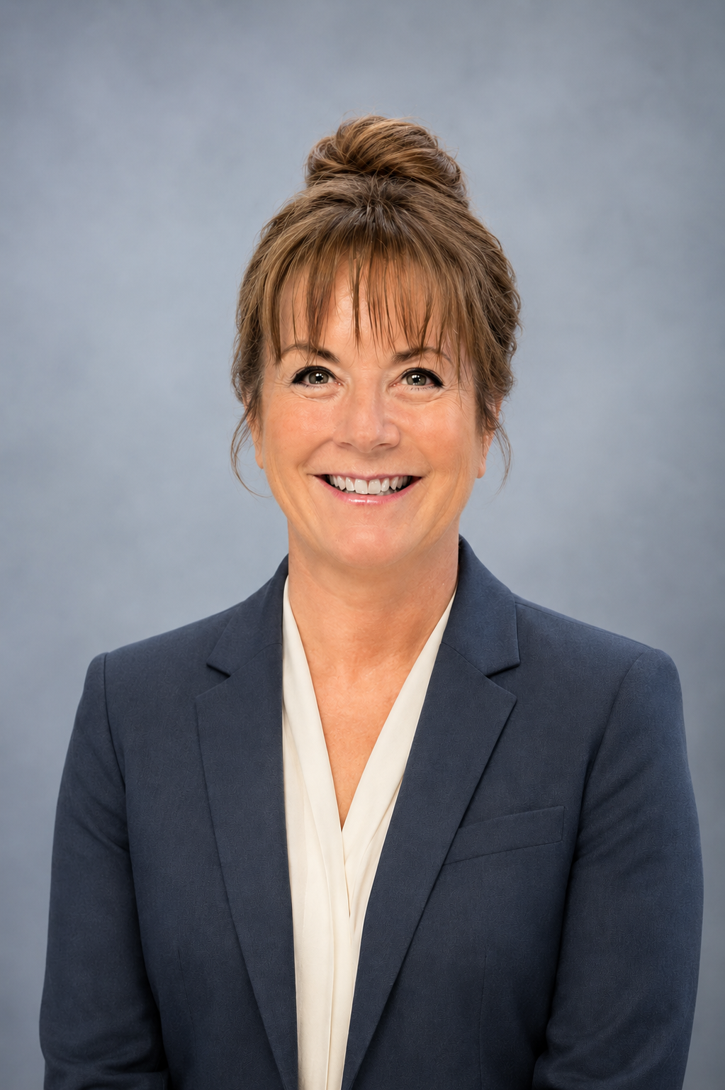 A woman with light skin, short brown hair in a bun, smiling, dressed in a dark blue blazer and white blouse, posing against a plain gray background.