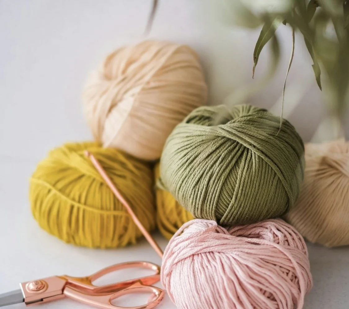 Colorful balls of yarn in pink, yellow, green, and beige with a pair of pink scissors on a white surface.