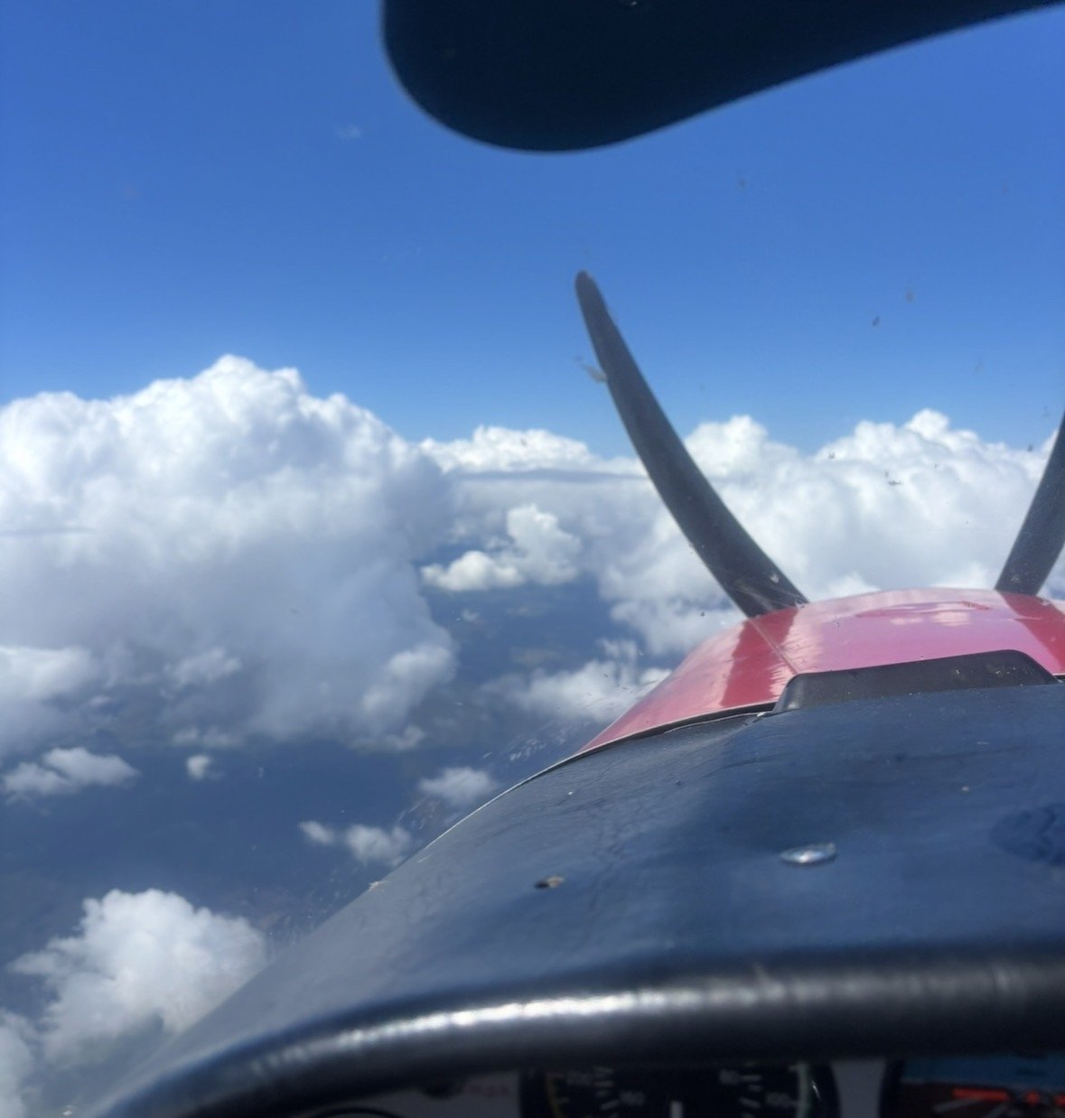Photo taken from inside a small airplane cockpit, showing the sky with clouds and the airplane's nose and wing.