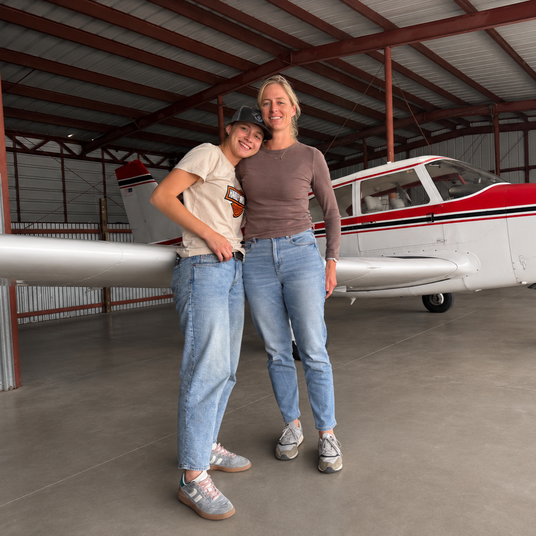 Two women standing inside an airplane hangar, smiling at the camera. One woman is wearing a Harley-Davidson t-shirt and a cap, the other is in a long-sleeve shirt. They are positioned in front of a small white and red airplane.