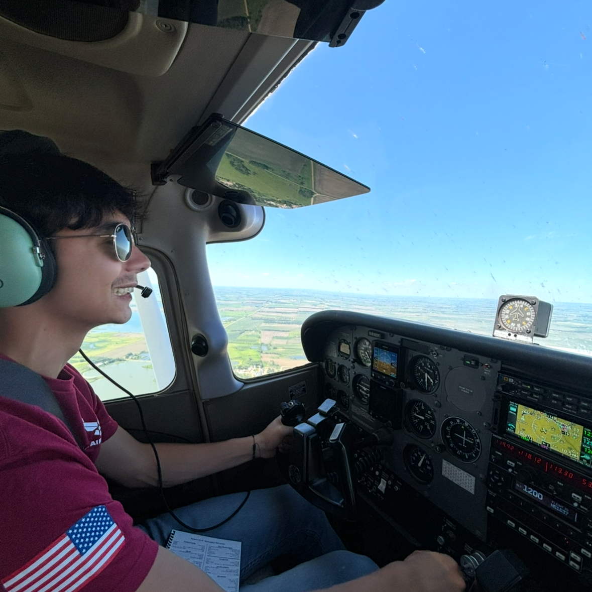 A young man flying a small airplane, wearing sunglasses and a headset, with a view of the landscape below through the cockpit window.