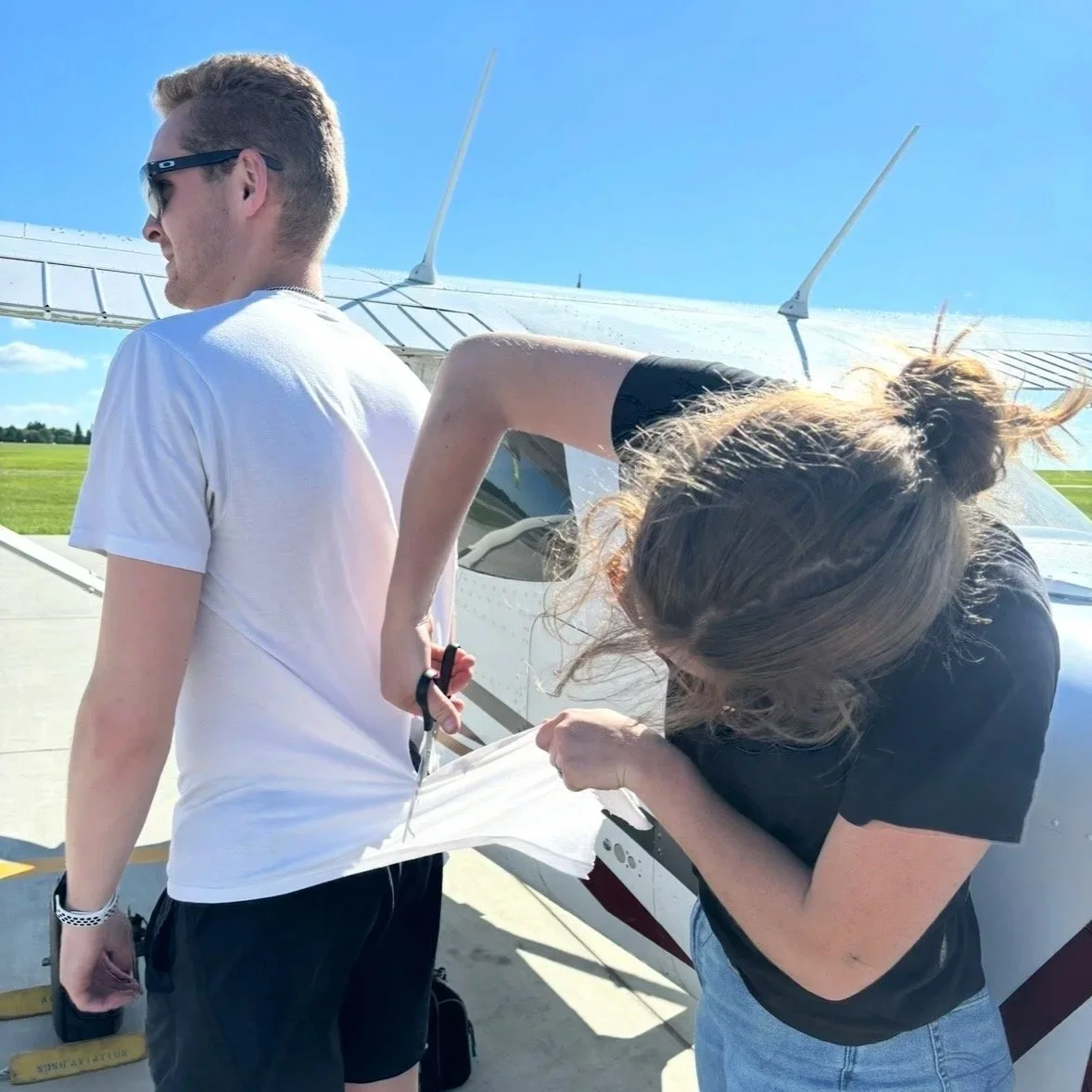 A woman with light brown hair in a bun cuts a mans t shirt symbolizing his first solo flight near a small airplane on a sunny day.