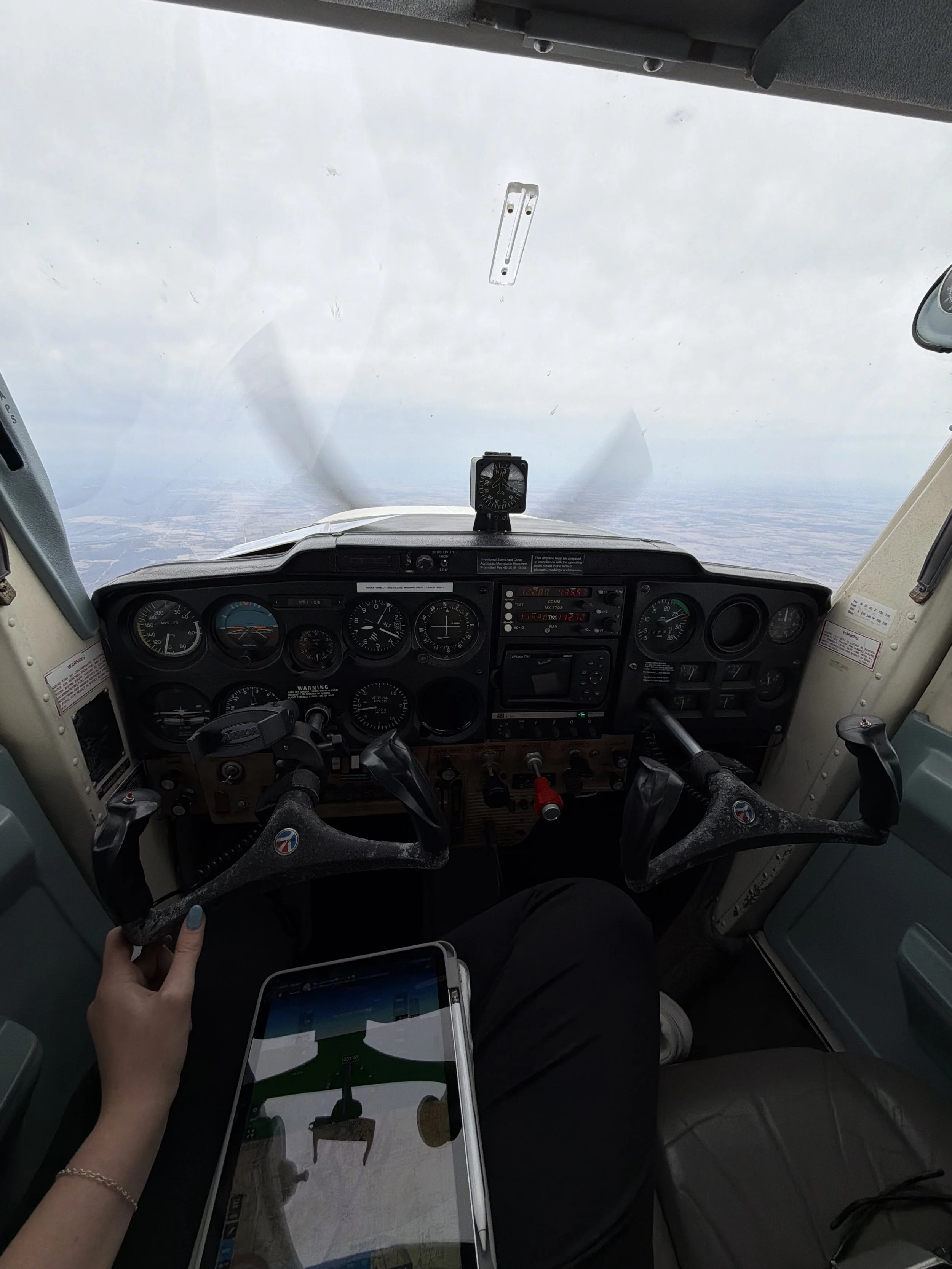 Pilot cockpit view showing airplane instruments, a hand holding a tablet device, and a partially visible person's leg with a smartphone. The view outside the cockpit shows cloudy skies and landscape below.