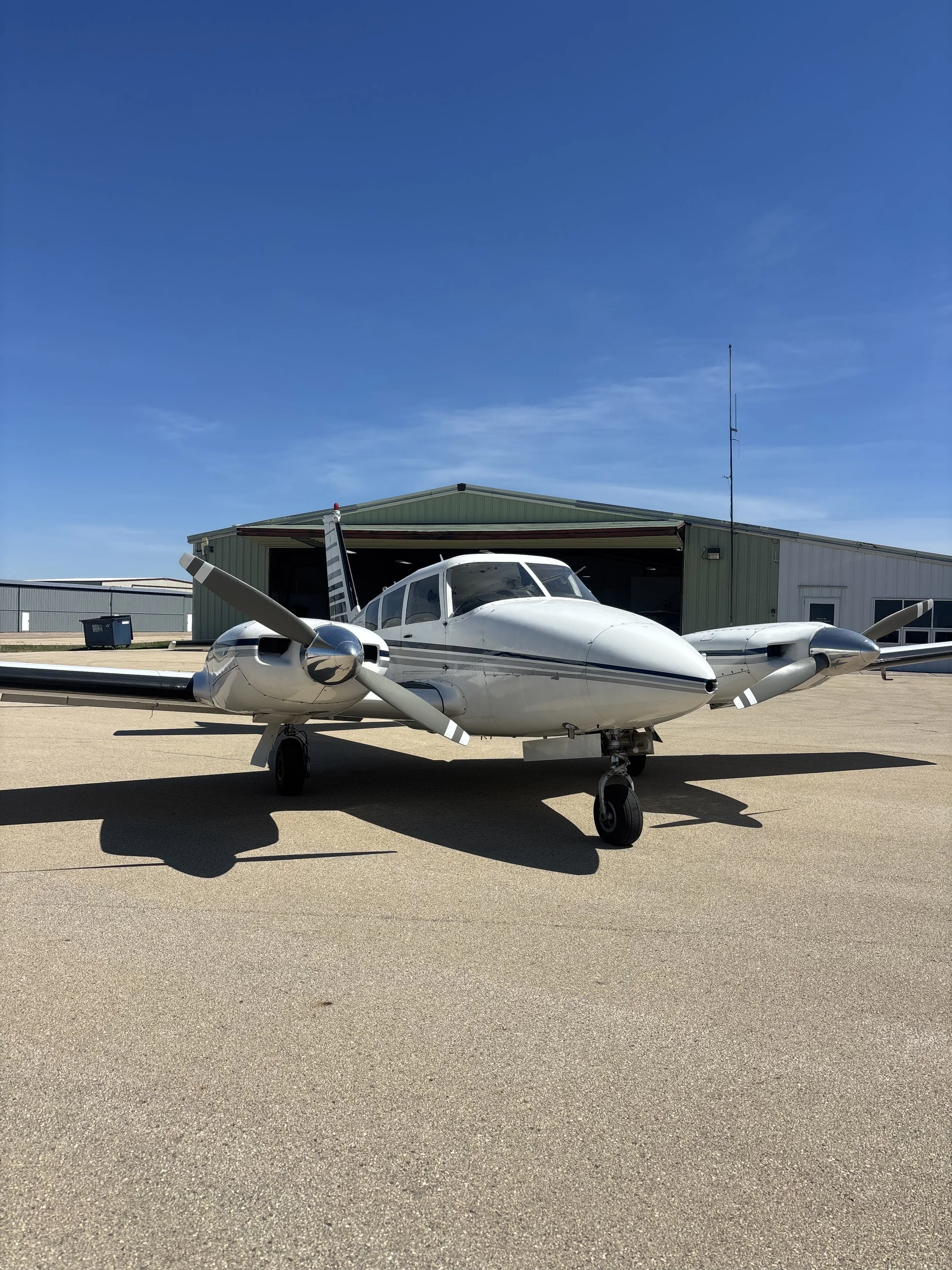 A small white aircraft with twin engines parked on a tarmac in front of a green hanger building under a clear blue sky.