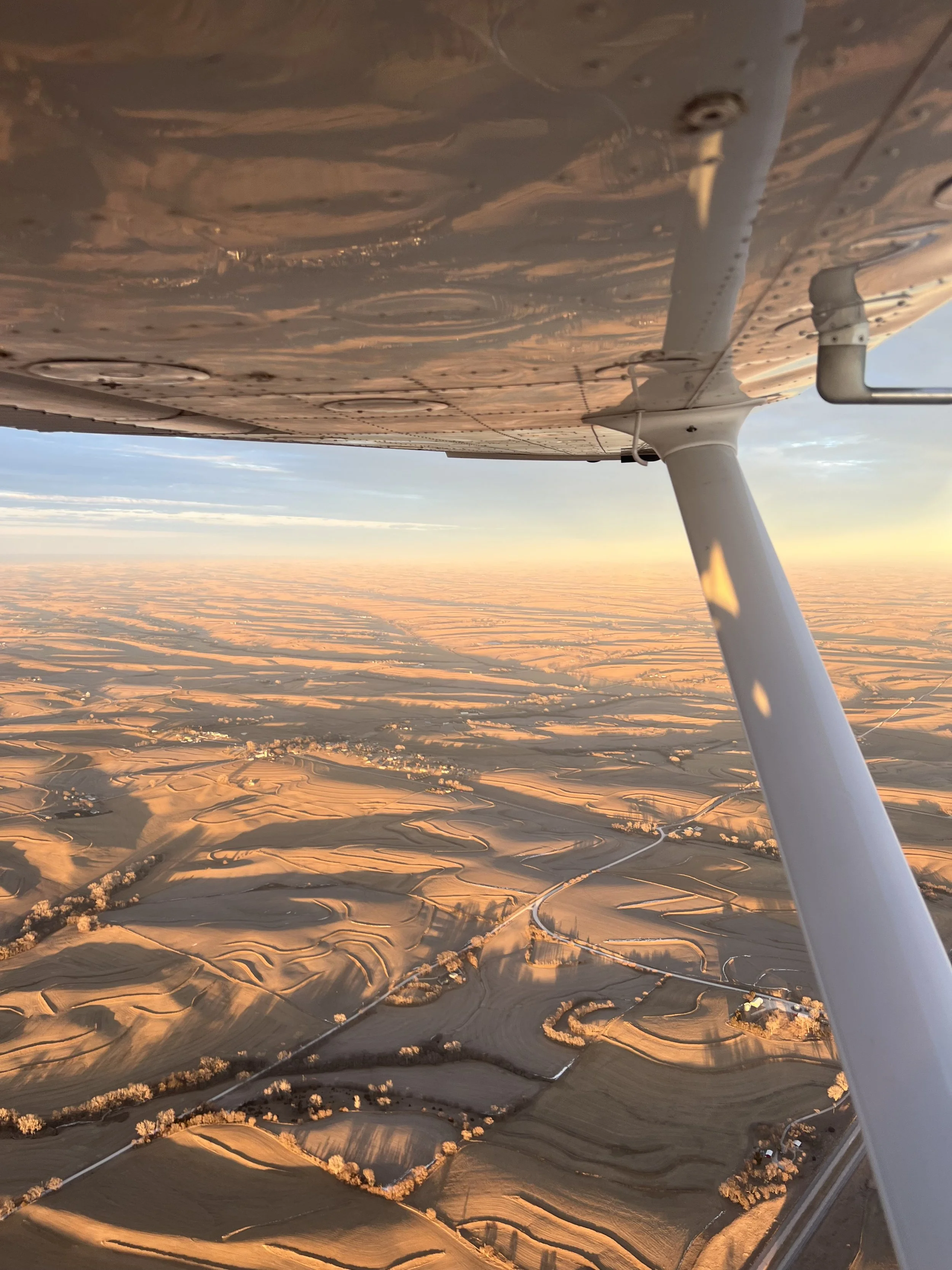 A view from an airplane window shows a landscape of fields and farmland during sunset, with the airplane's wing in the foreground.