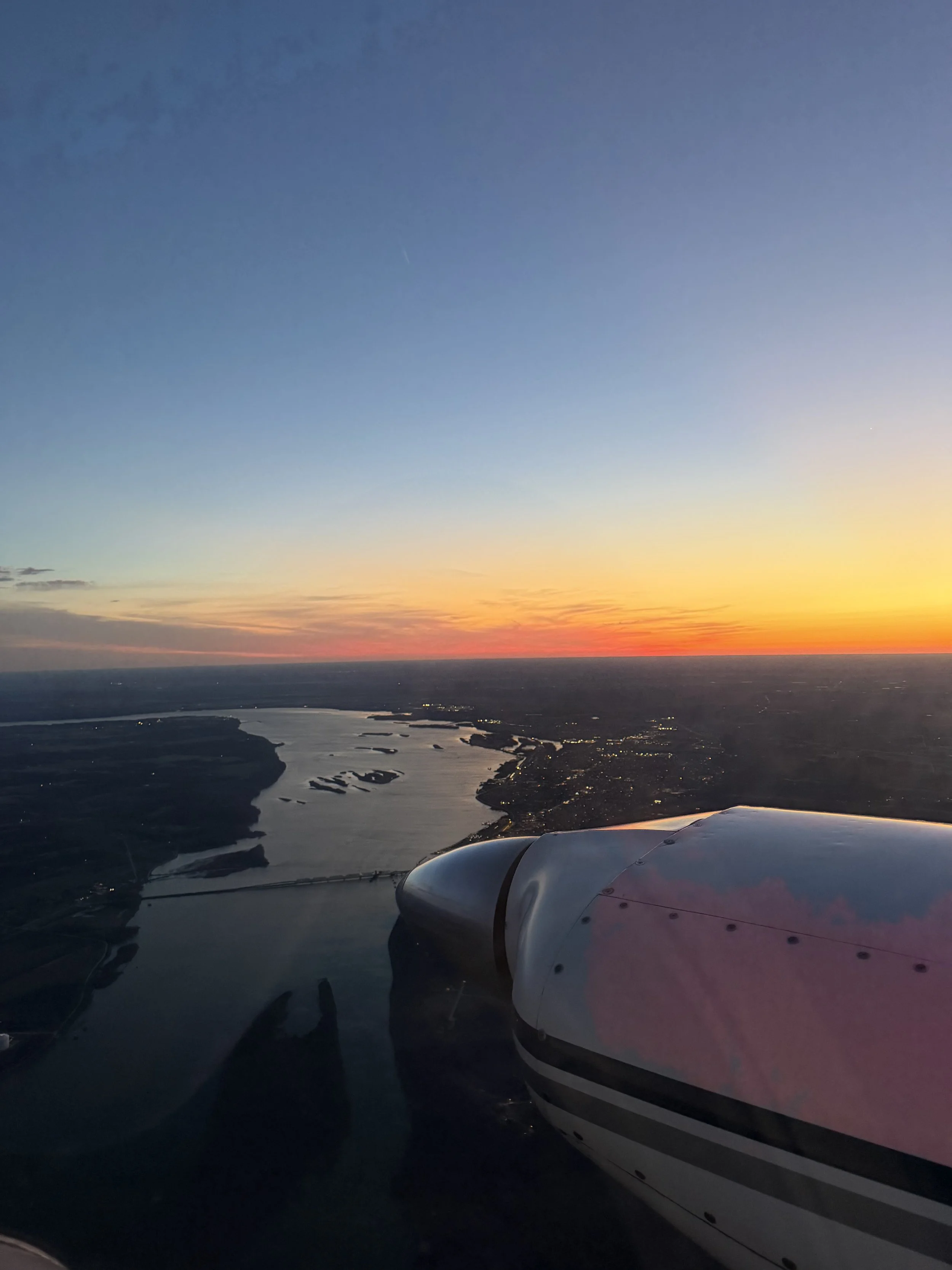 View from an airplane window showing a sunset over a river with land and water bodies, with the airplane engine visible in the bottom right corner.