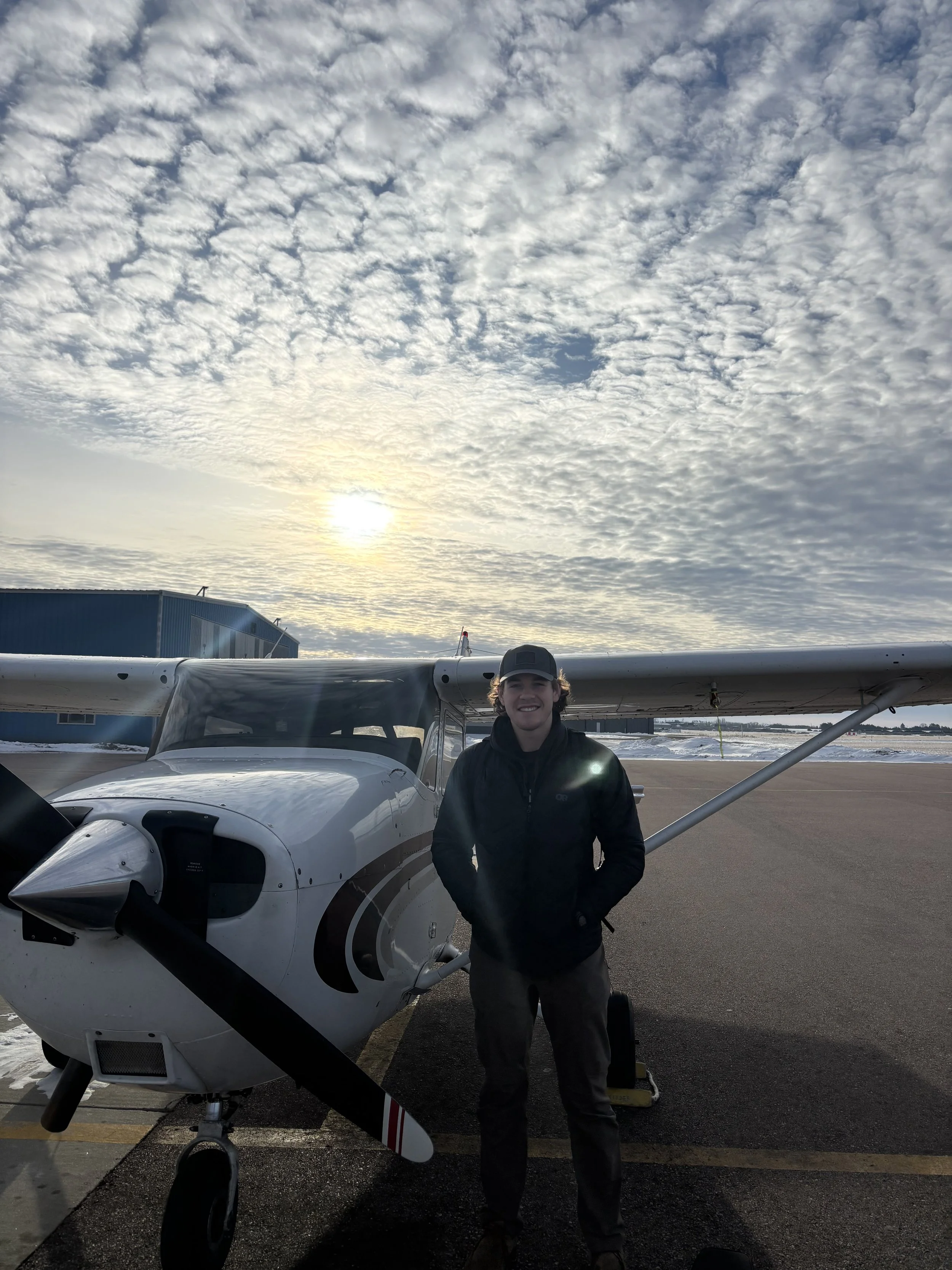 A young man wearing a black jacket and cap standing next to a small white aircraft on a tarmac, with a cloudy sky and sun in the background.