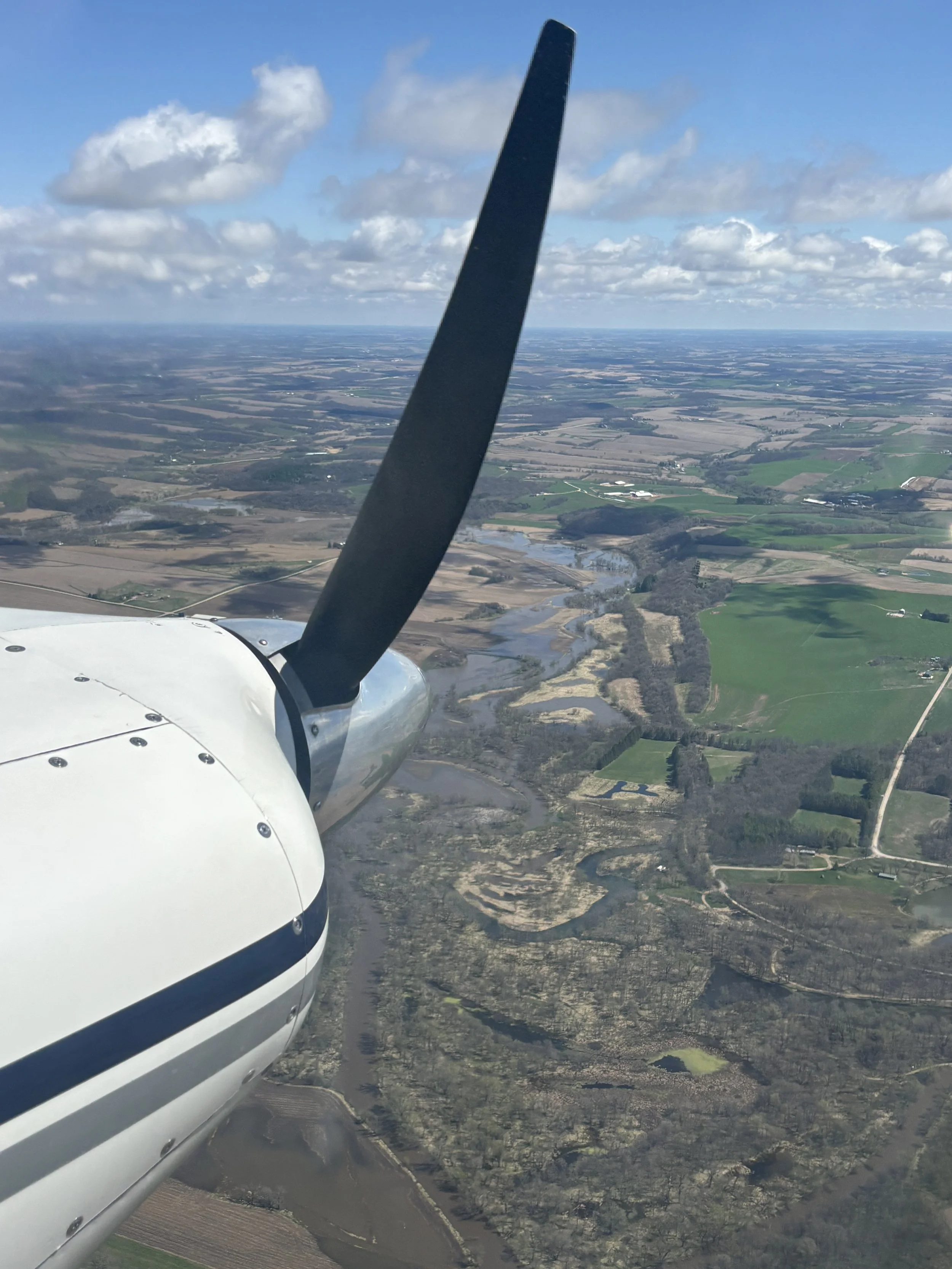 A view from a small airplane showing land, water, and fields with clouds in the sky.