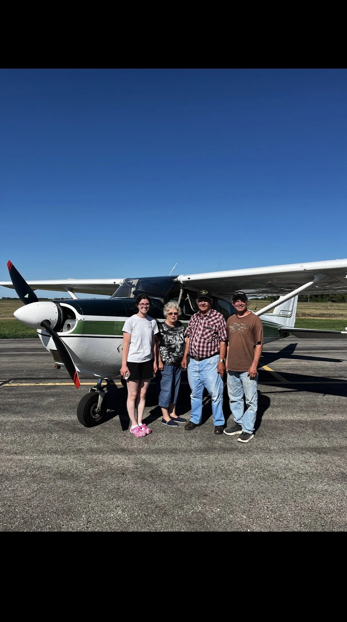 Four people standing in front of a small airplane on a runway at an airport under a clear blue sky.