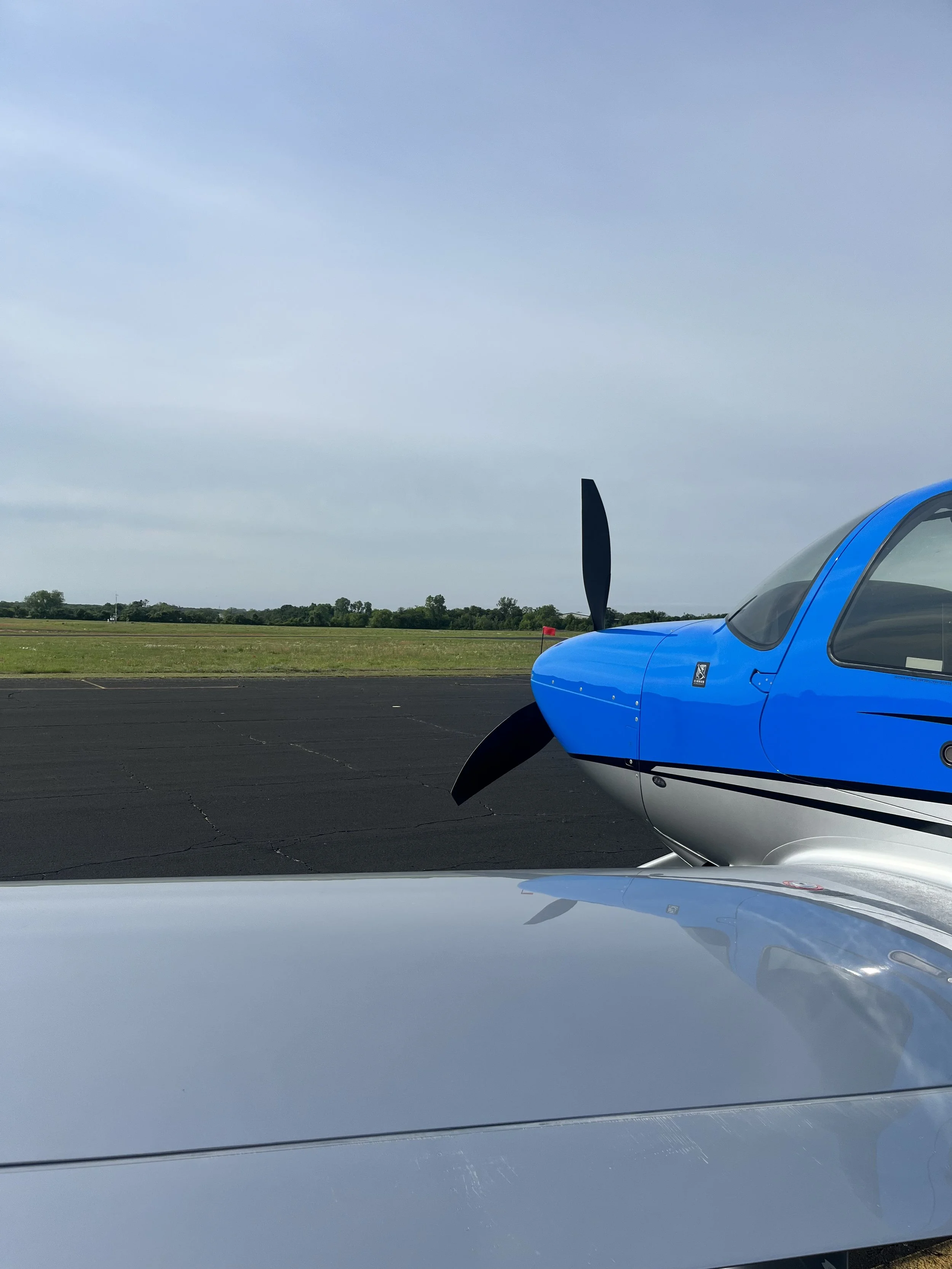 A blue airplane parked on a runway with a grassy field and trees in the background under a partly cloudy sky.